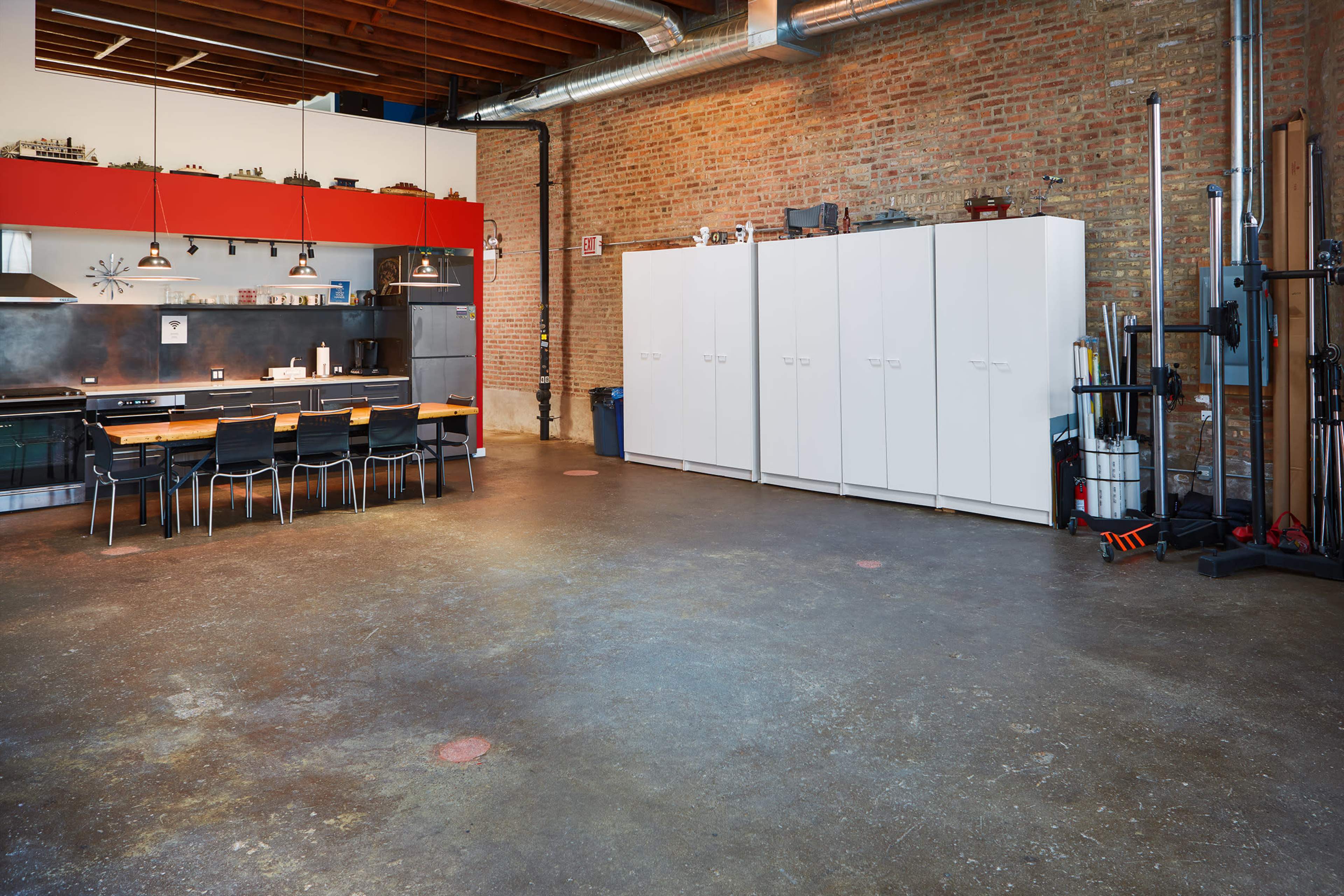 A spacious industrial-style kitchen with a wooden dining table, black chairs, and white cabinets against a brick wall.
