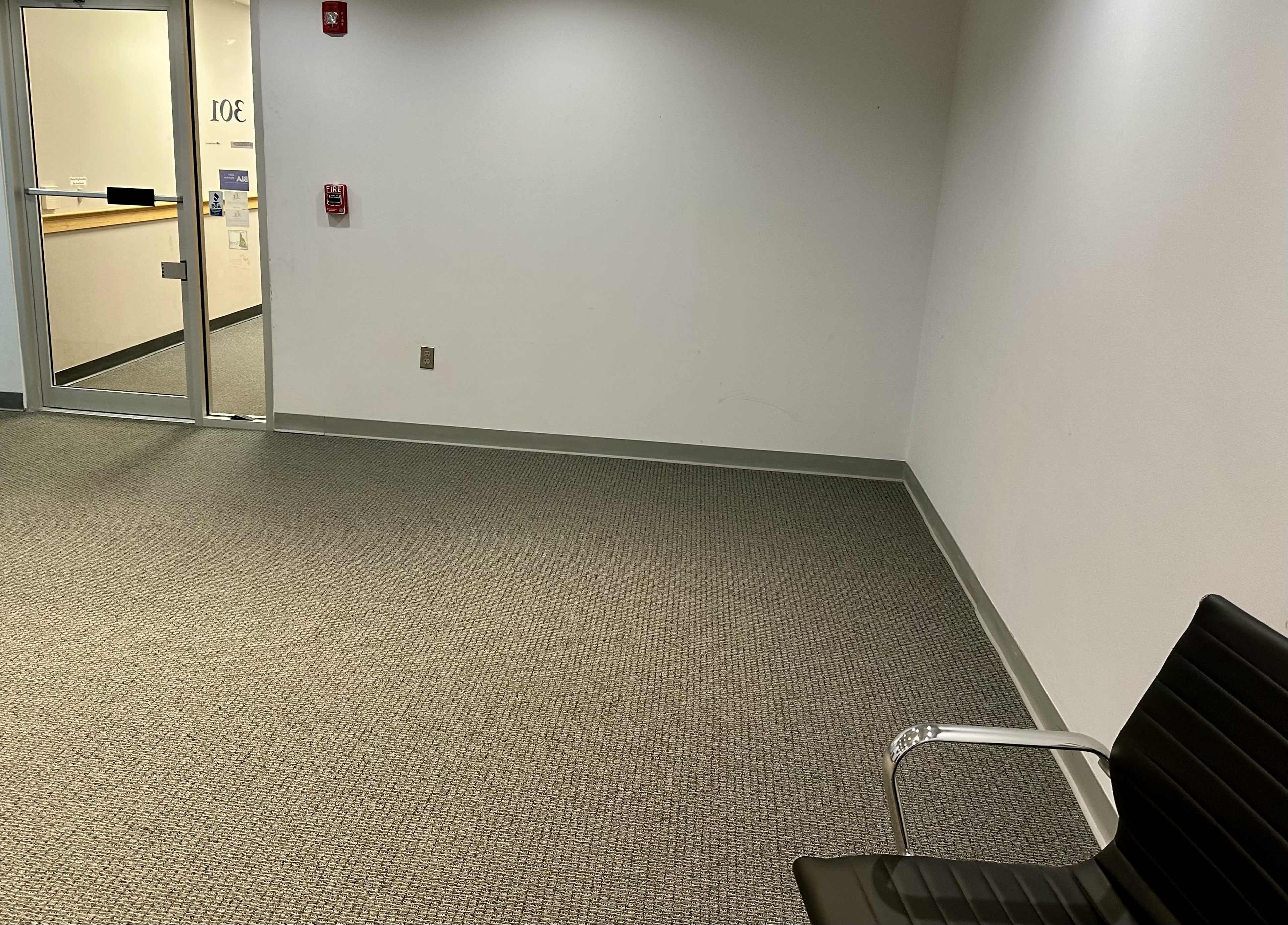 The image shows a sparsely furnished room with a single black chair and a glass door leading to another area.