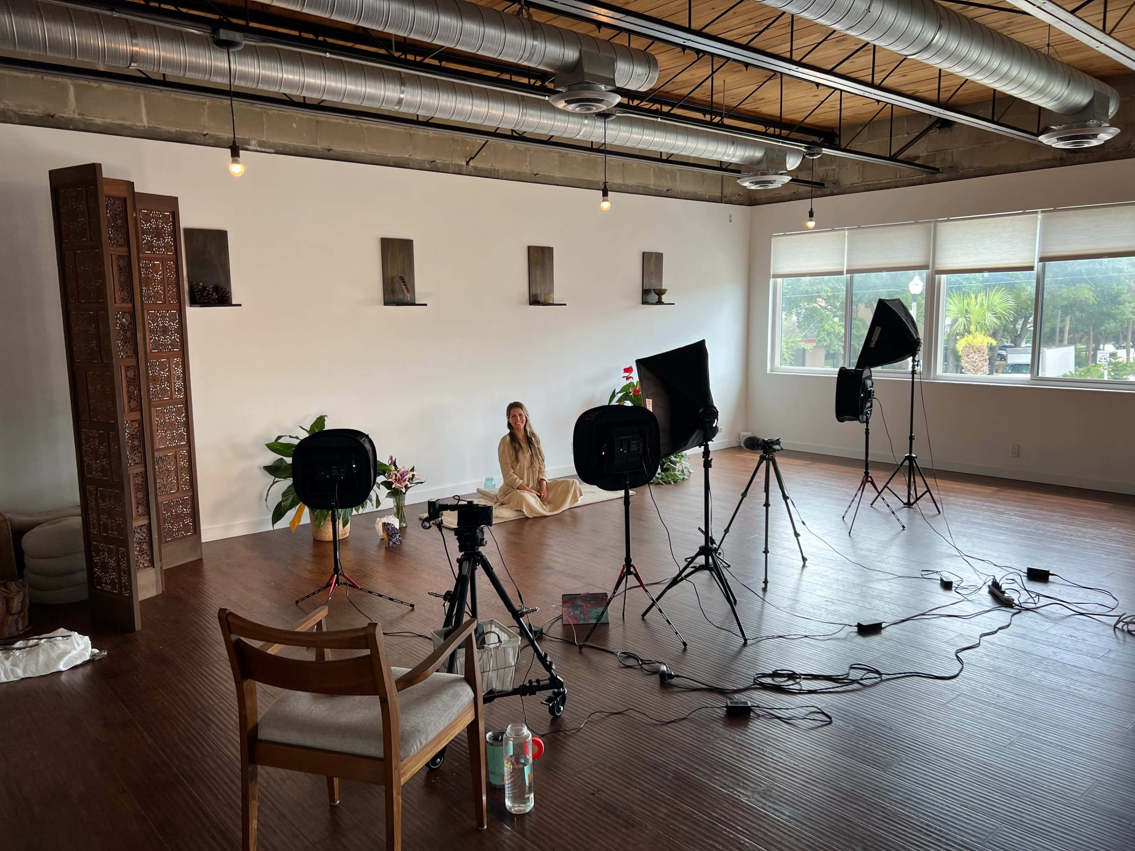 A spacious room set up for filming, featuring lights and cameras focused on a seated person amidst decorative plants and wall shelves.