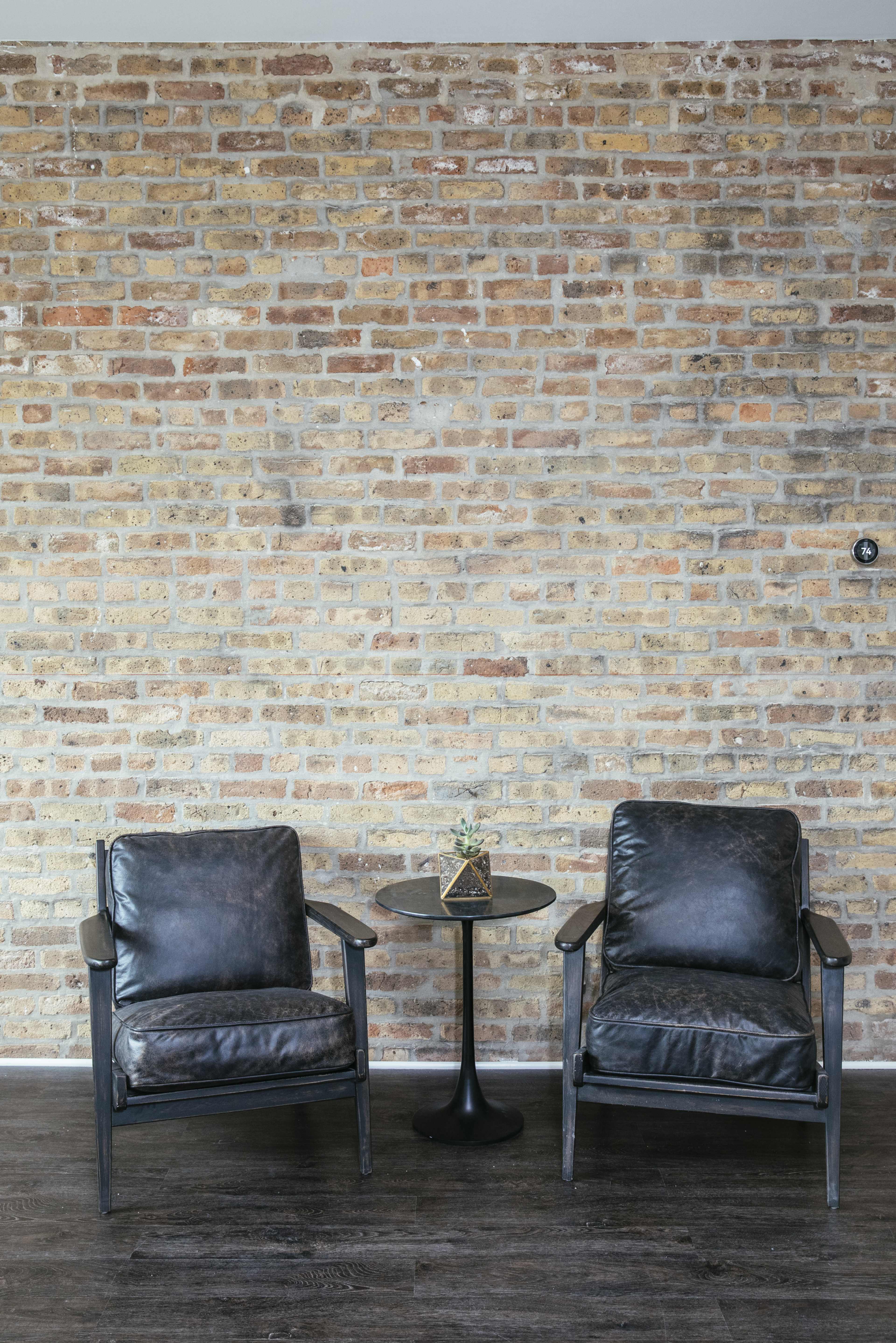 Two black chairs are positioned on either side of a small round table against a brick wall.