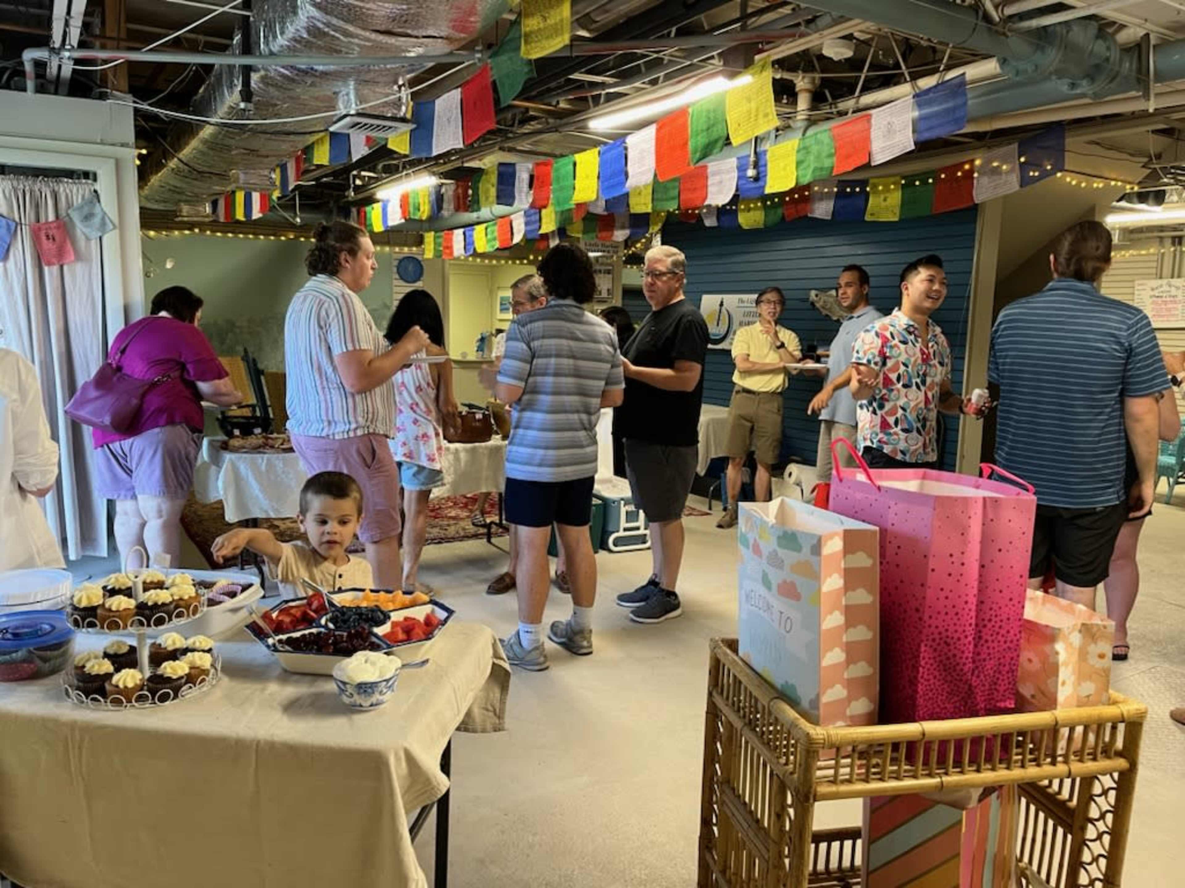 A group of people socialize in a decorated basement space filled with tables of food, colorful banners overhead, and large gift bags on display.