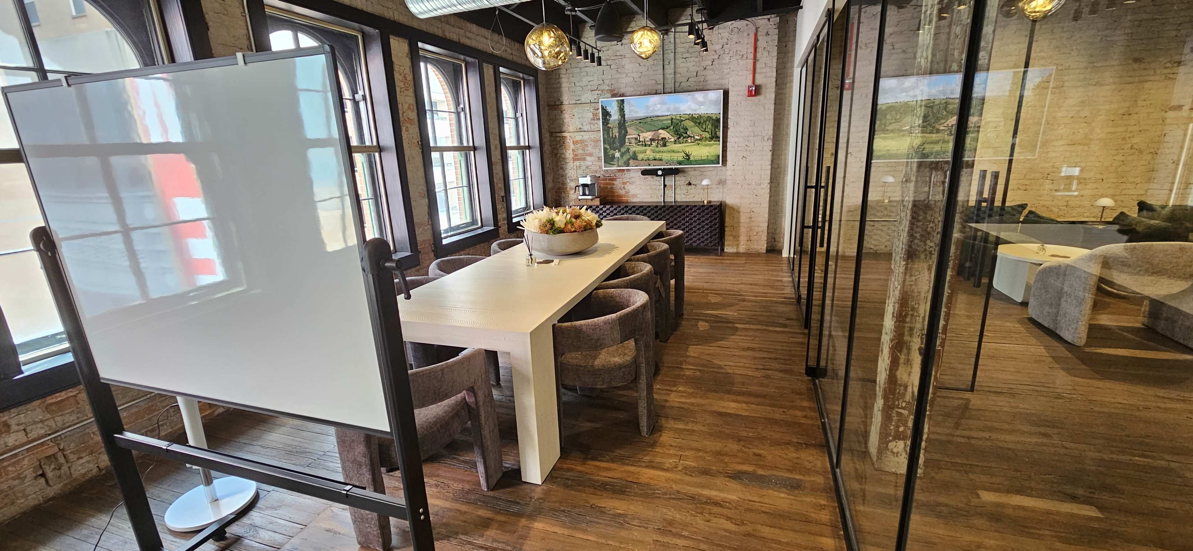 A modern conference room with a white dining table surrounded by brown chairs, a large window, and a television displaying a landscape image.