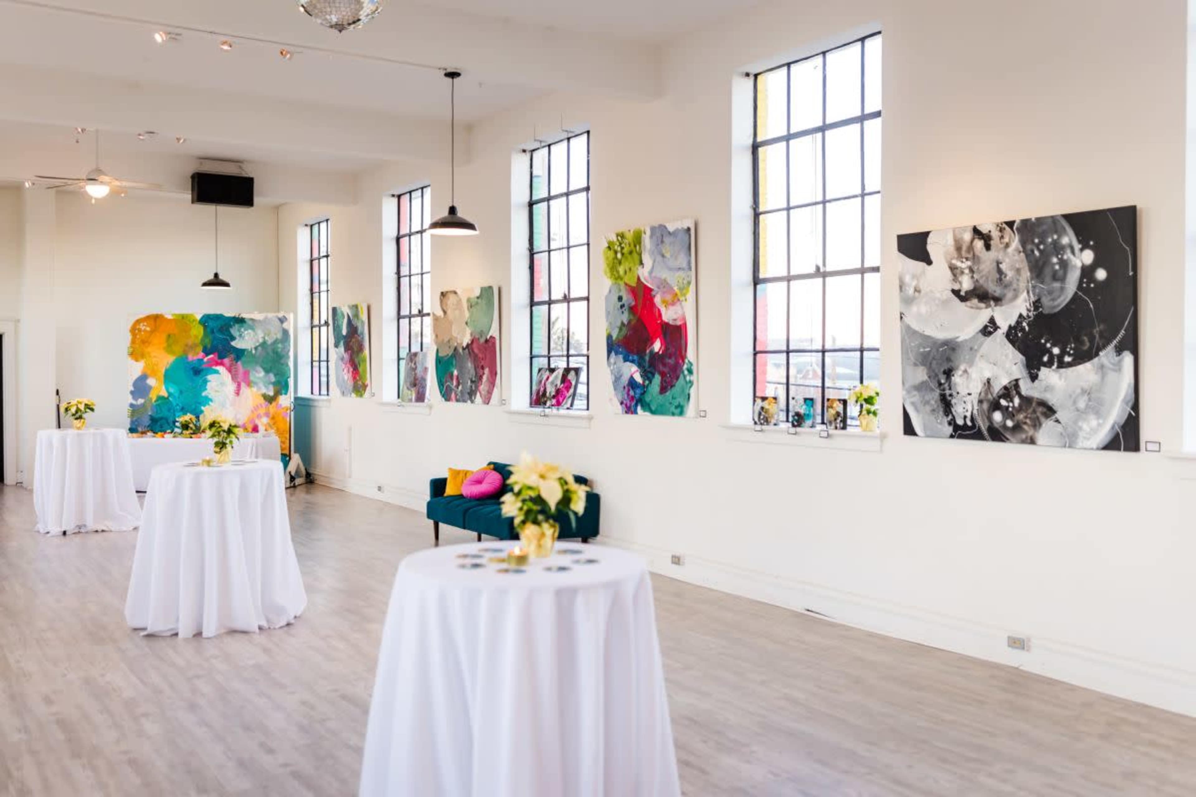 The image shows a well-lit gallery space featuring tables covered with white cloths, a green sofa, and large abstract paintings mounted on the walls.