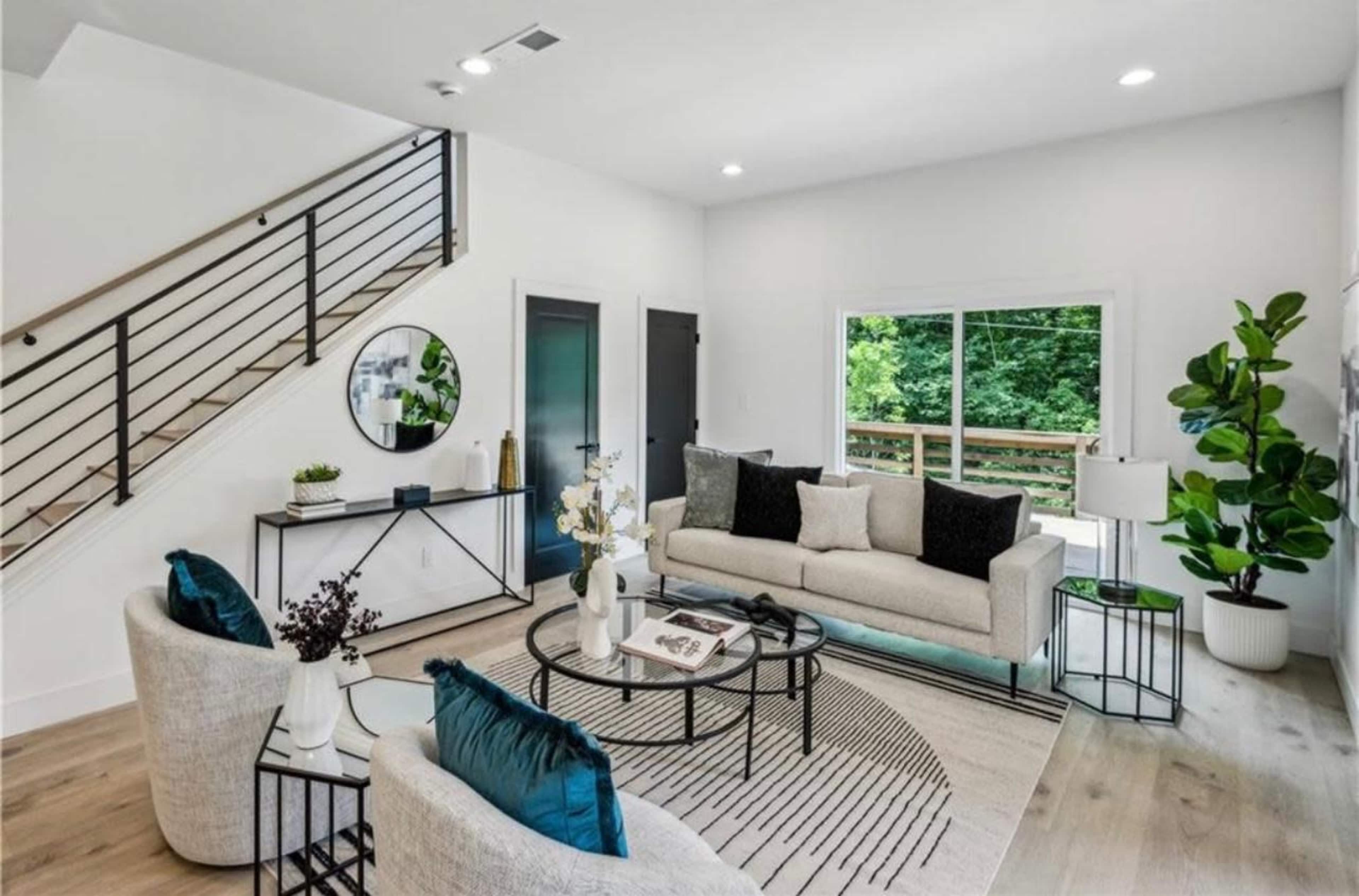 A modern living room features a light-colored sofa, two armchairs, a round coffee table, and large windows with a view of greenery outside.