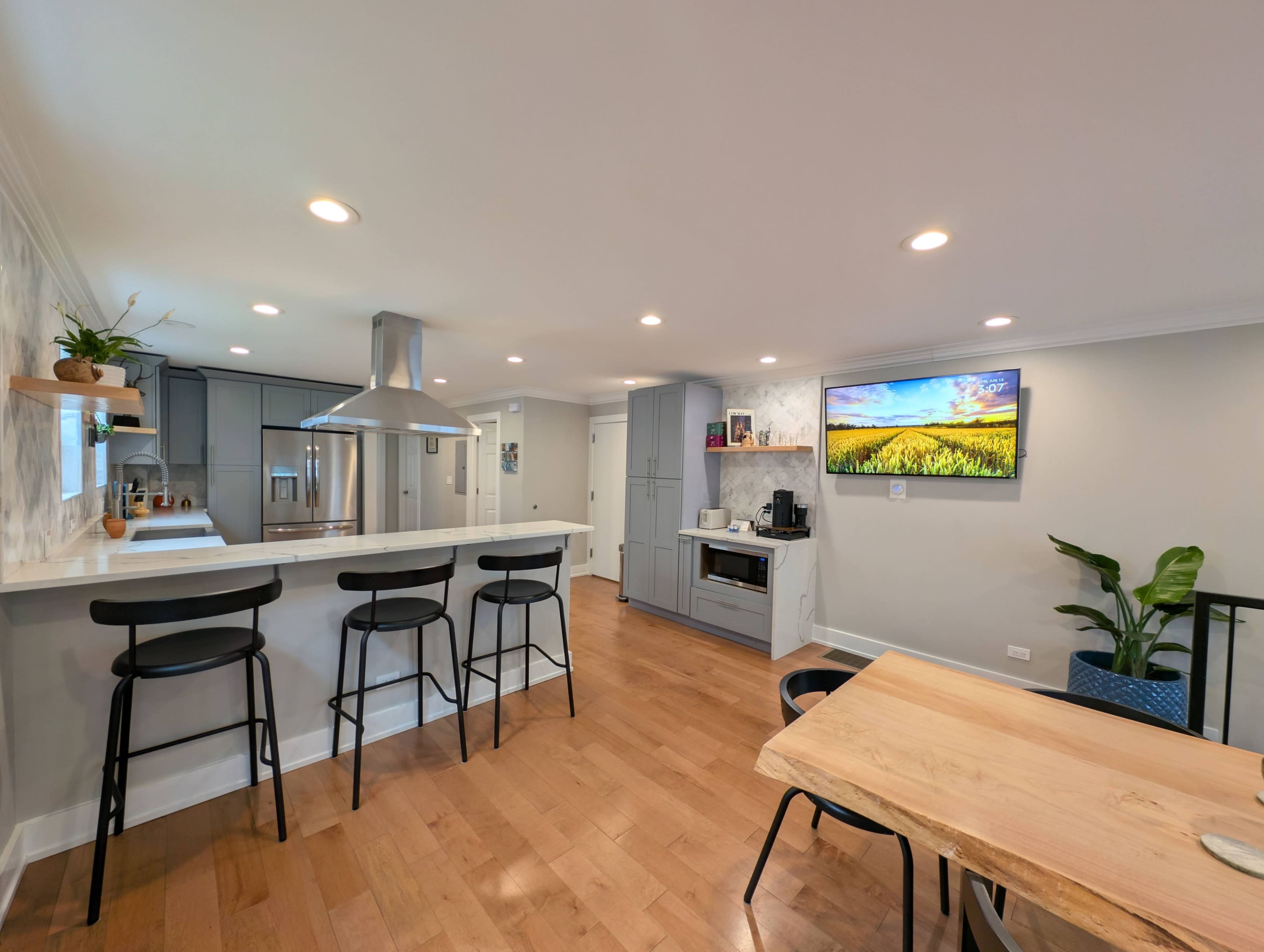 A modern kitchen features gray cabinetry, a central island with bar seating, a dining table, and a large wall-mounted television.