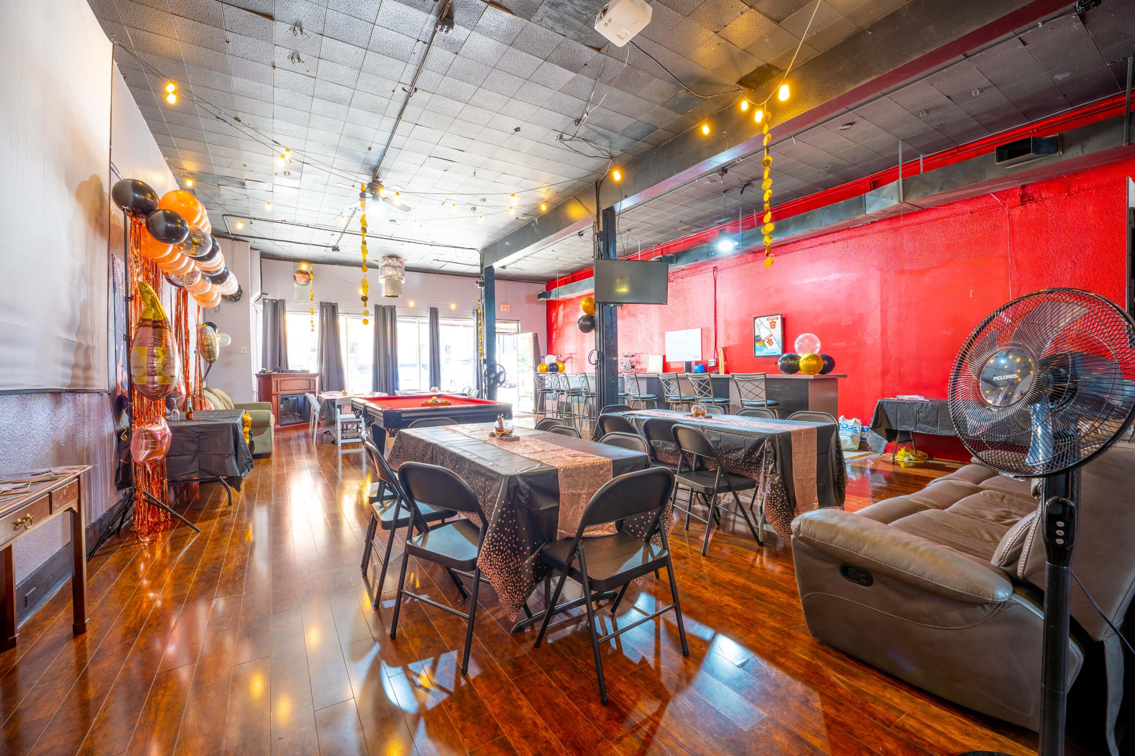 The image shows a spacious event room decorated with tables, chairs, and string lights, featuring a pool table and a bar area.