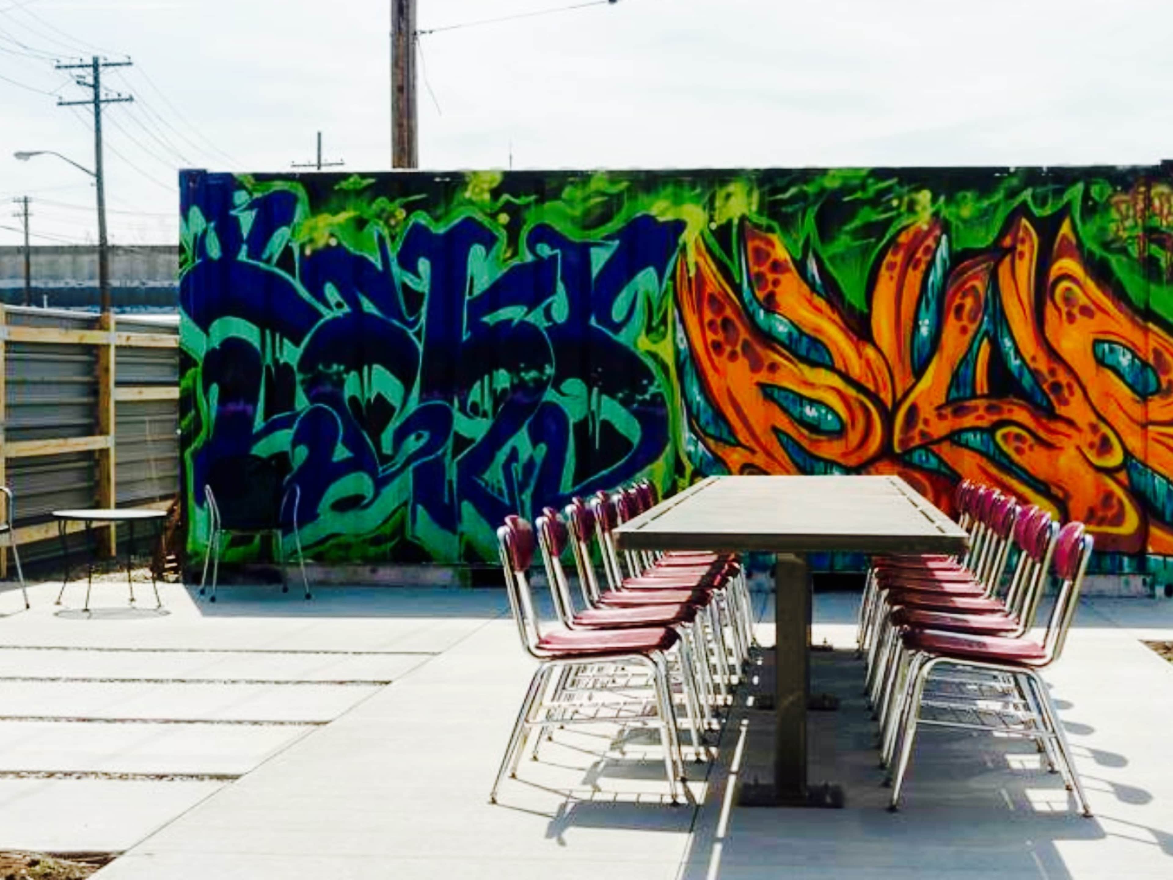 An outdoor space features a long table surrounded by chairs against a vibrant graffiti wall.