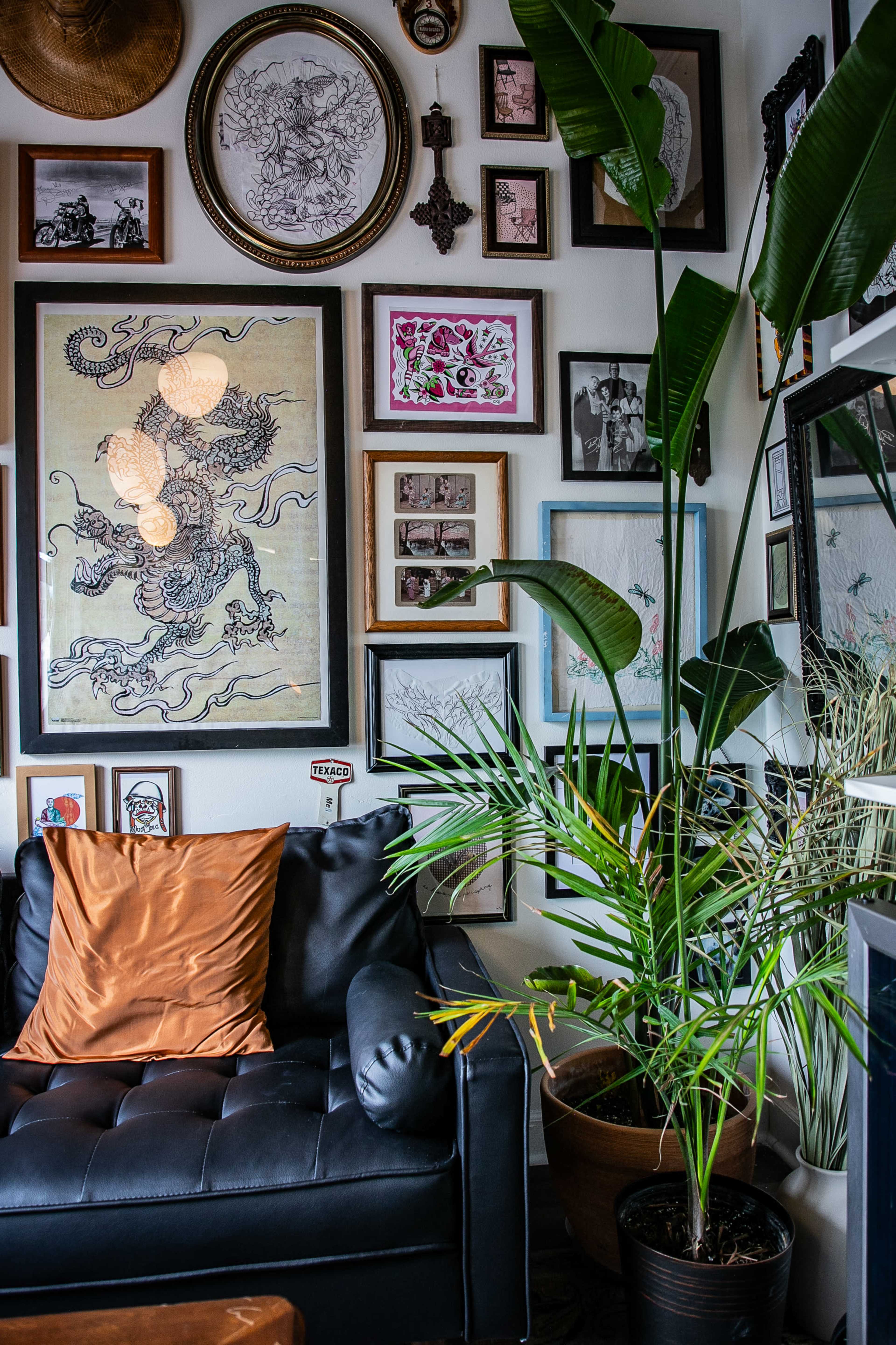 The image shows a black leather couch in a corner of a room adorned with various framed artworks and photographs on the walls, along with potted plants nearby.