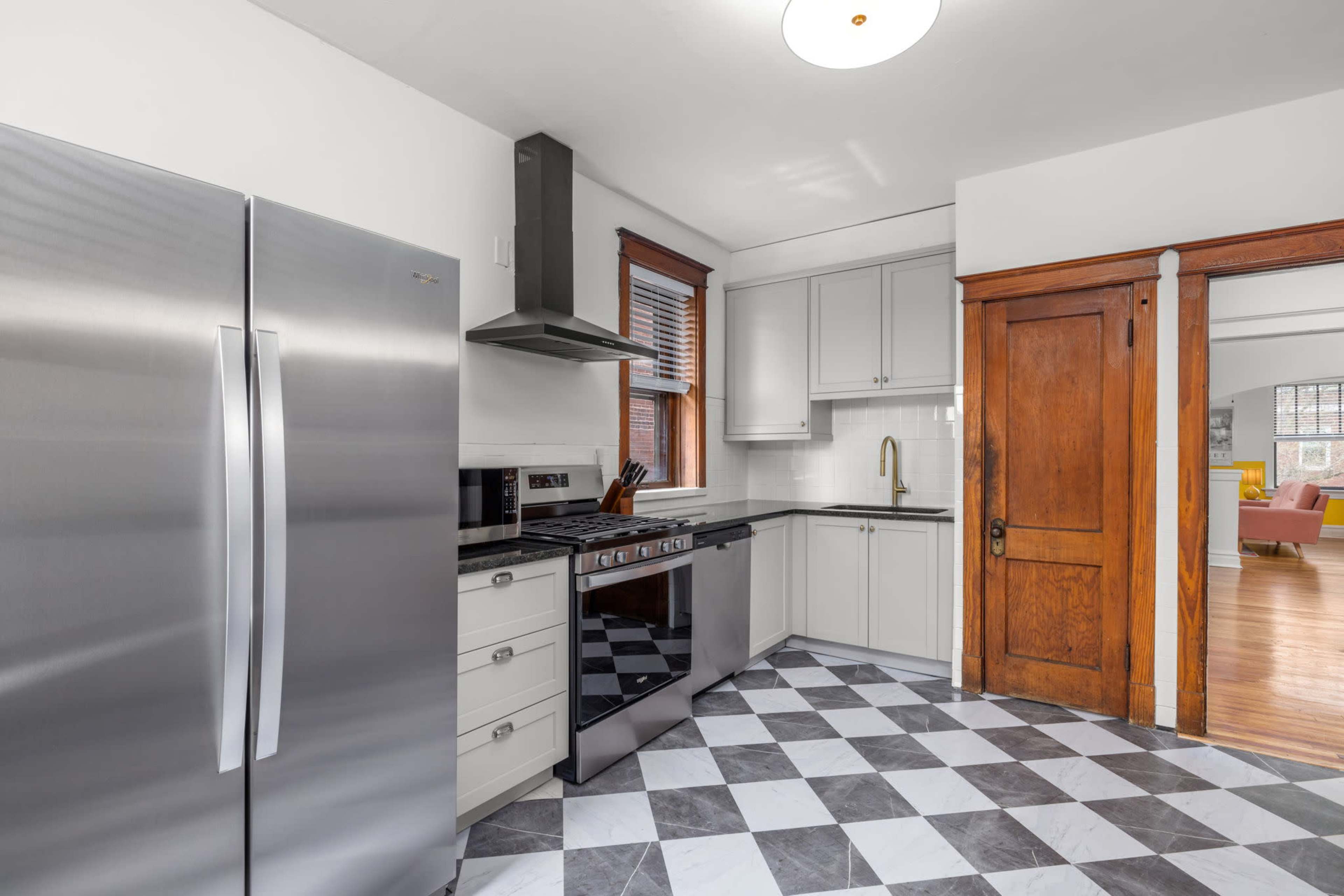 The image shows a modern kitchen with stainless steel appliances, gray and white checkered flooring, and wooden doorways leading into another room.