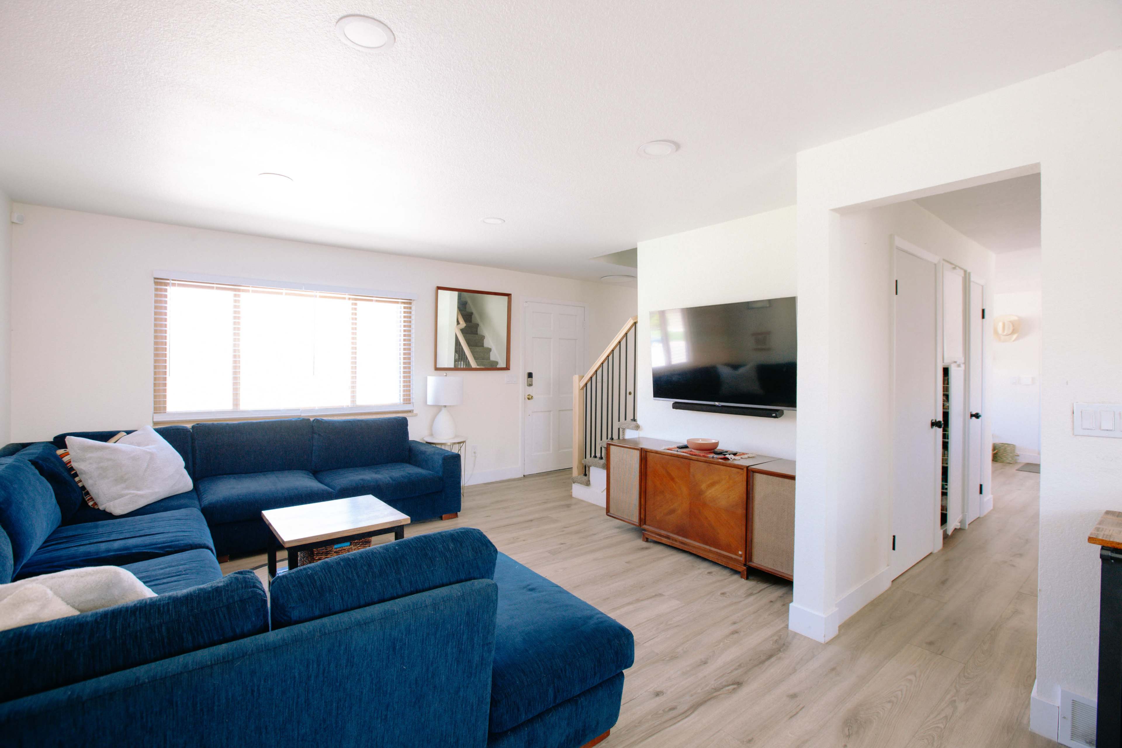 A modern living room featuring a blue sectional sofa, a wooden coffee table, a wall-mounted TV, and natural light coming through a window.