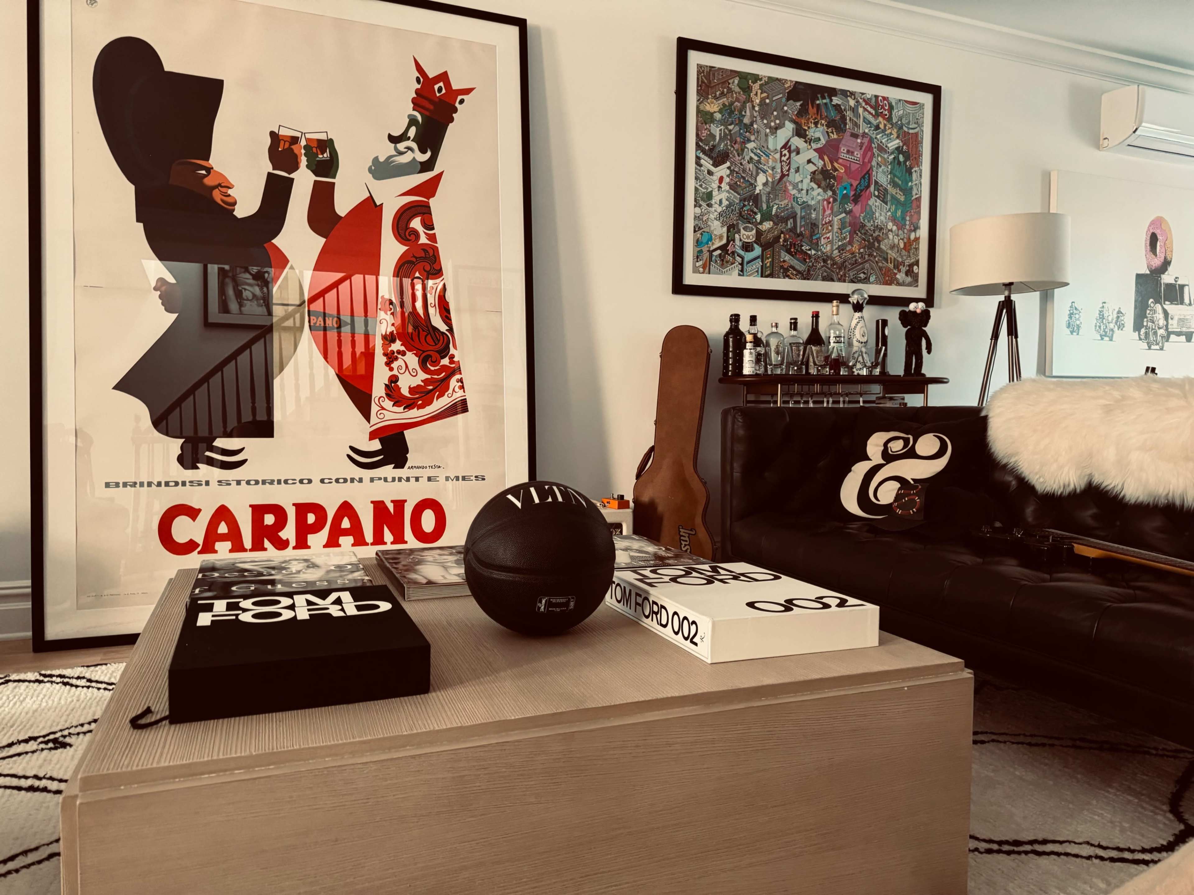 A stylish living room with a large vintage poster, a coffee table adorned with books and a basketball, and a black leather sofa with decorative cushions.