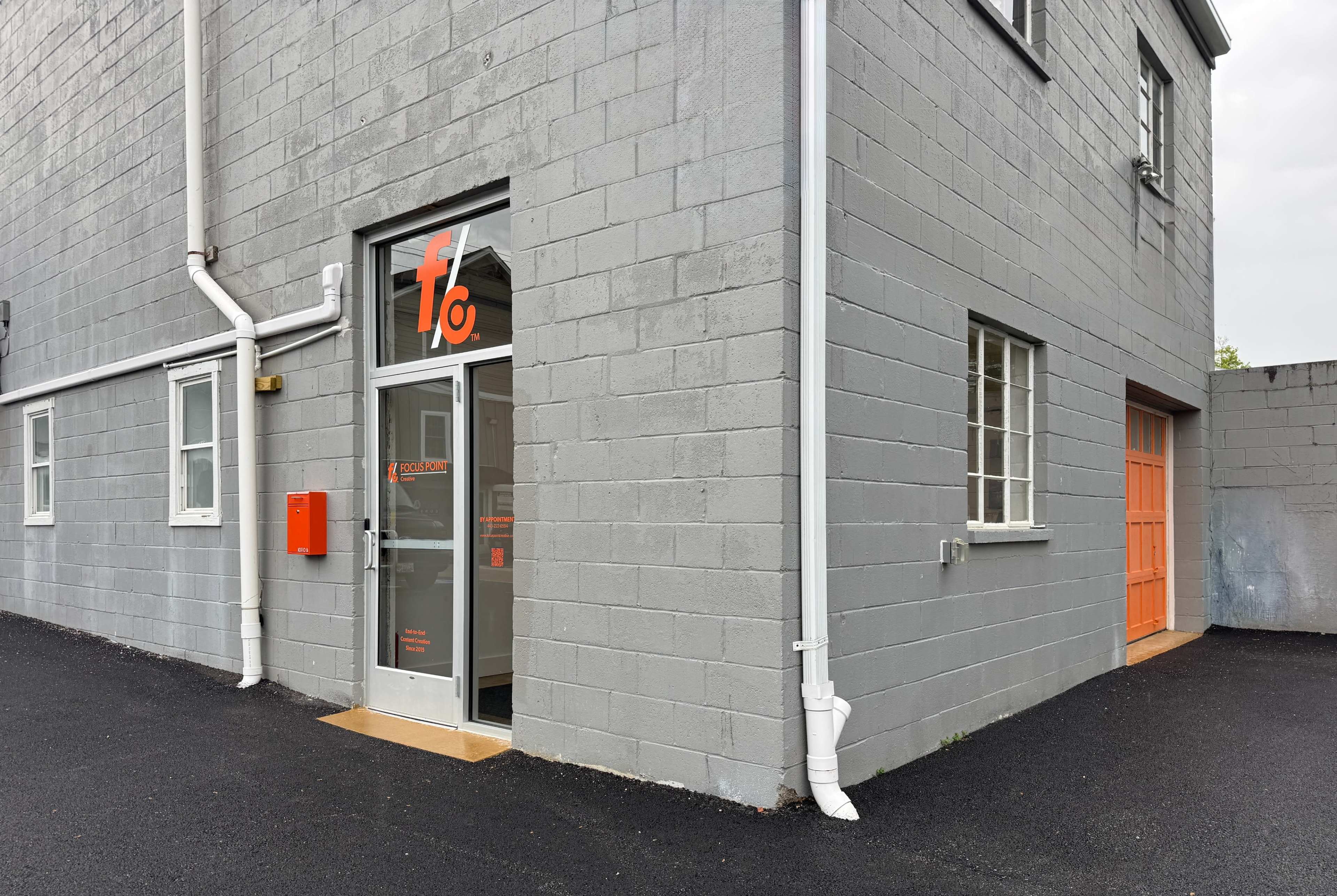 The image shows the entrance of a gray building with an orange accent door and a large glass door displaying the company logo.
