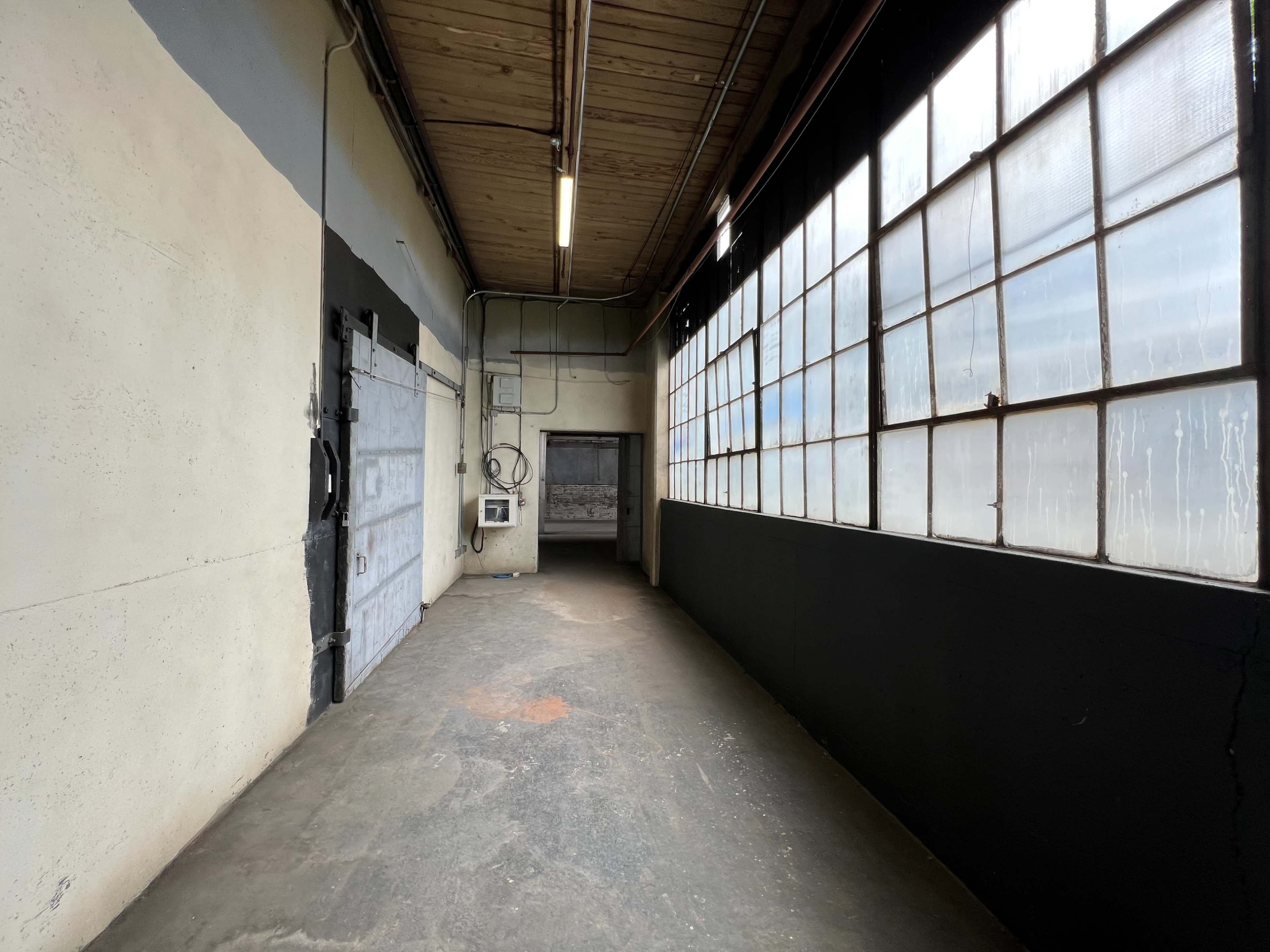 A long, narrow hallway with a concrete floor, metal doors, and large windows along one side.