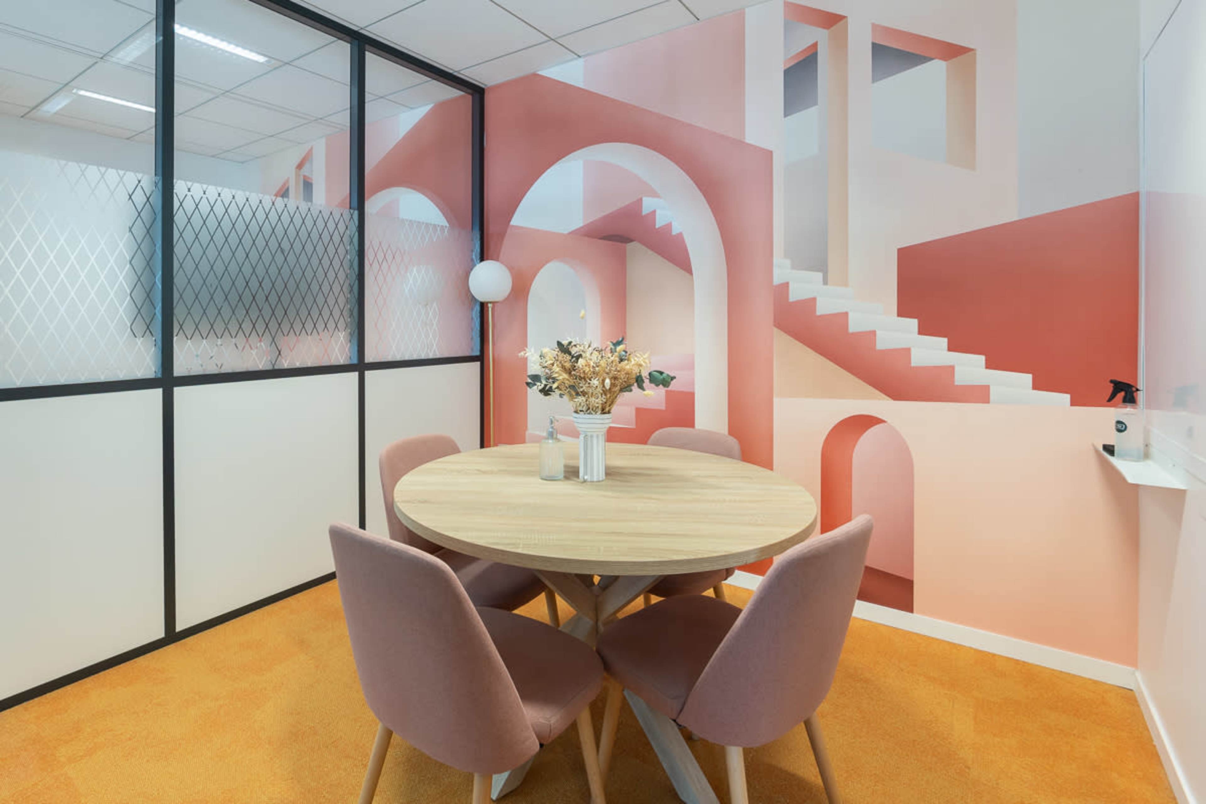 A modern meeting room features a round wooden table surrounded by pink chairs, with a colorful mural depicting stairs and arches on the wall.