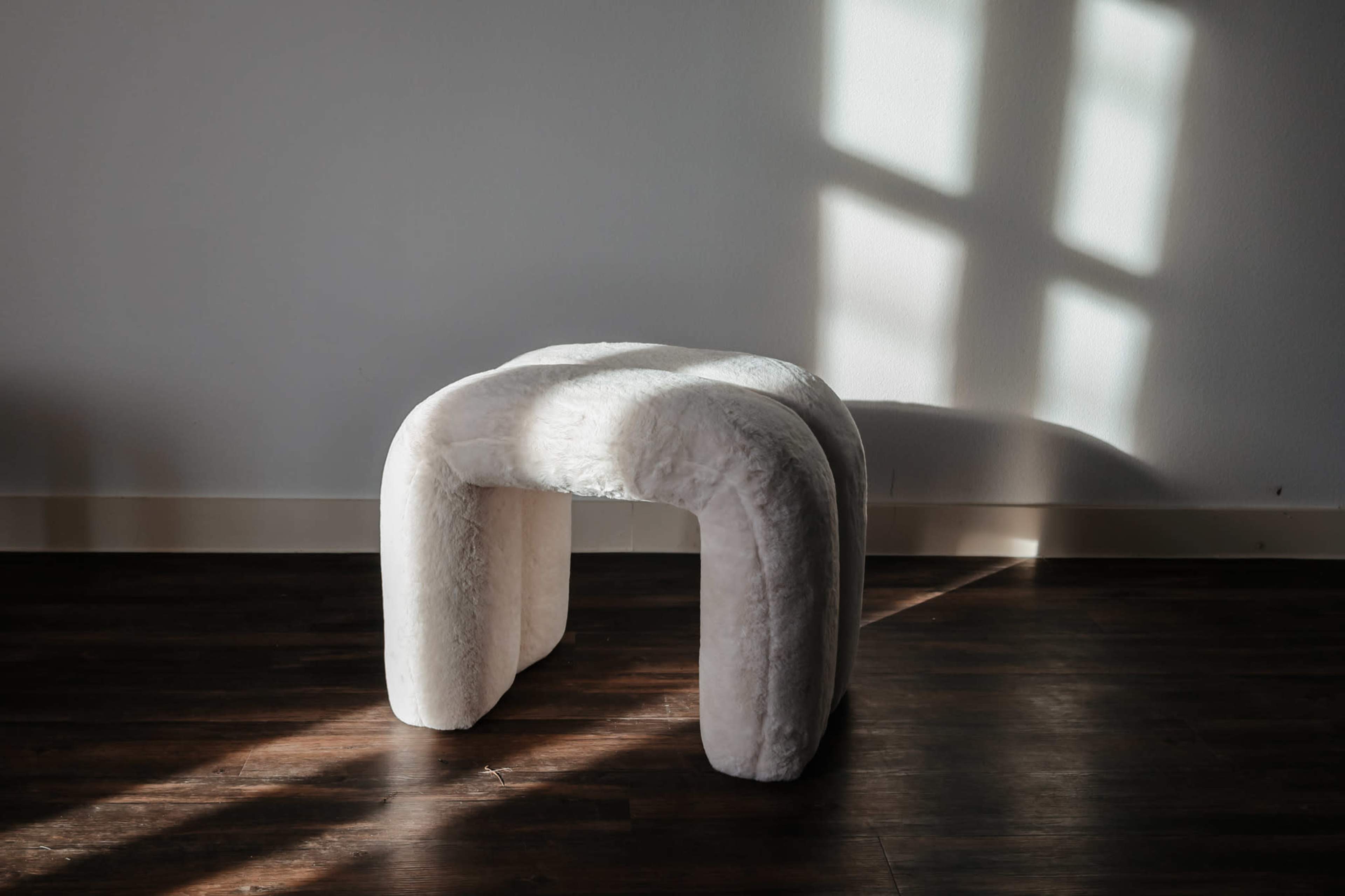 A white, upholstered stool with a curved design is positioned on a wooden floor under natural light.