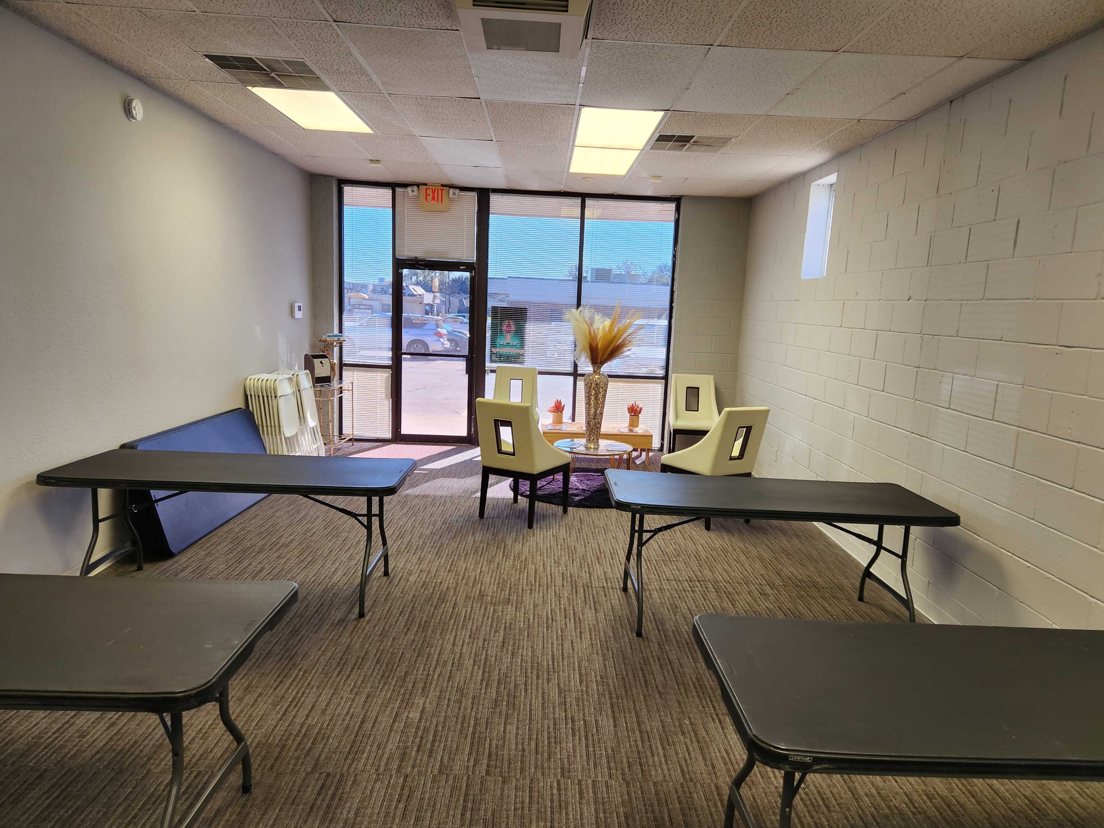 A room with several black tables arranged in a semi-circle, a seating area with chairs and a small table, and a large window letting in natural light.