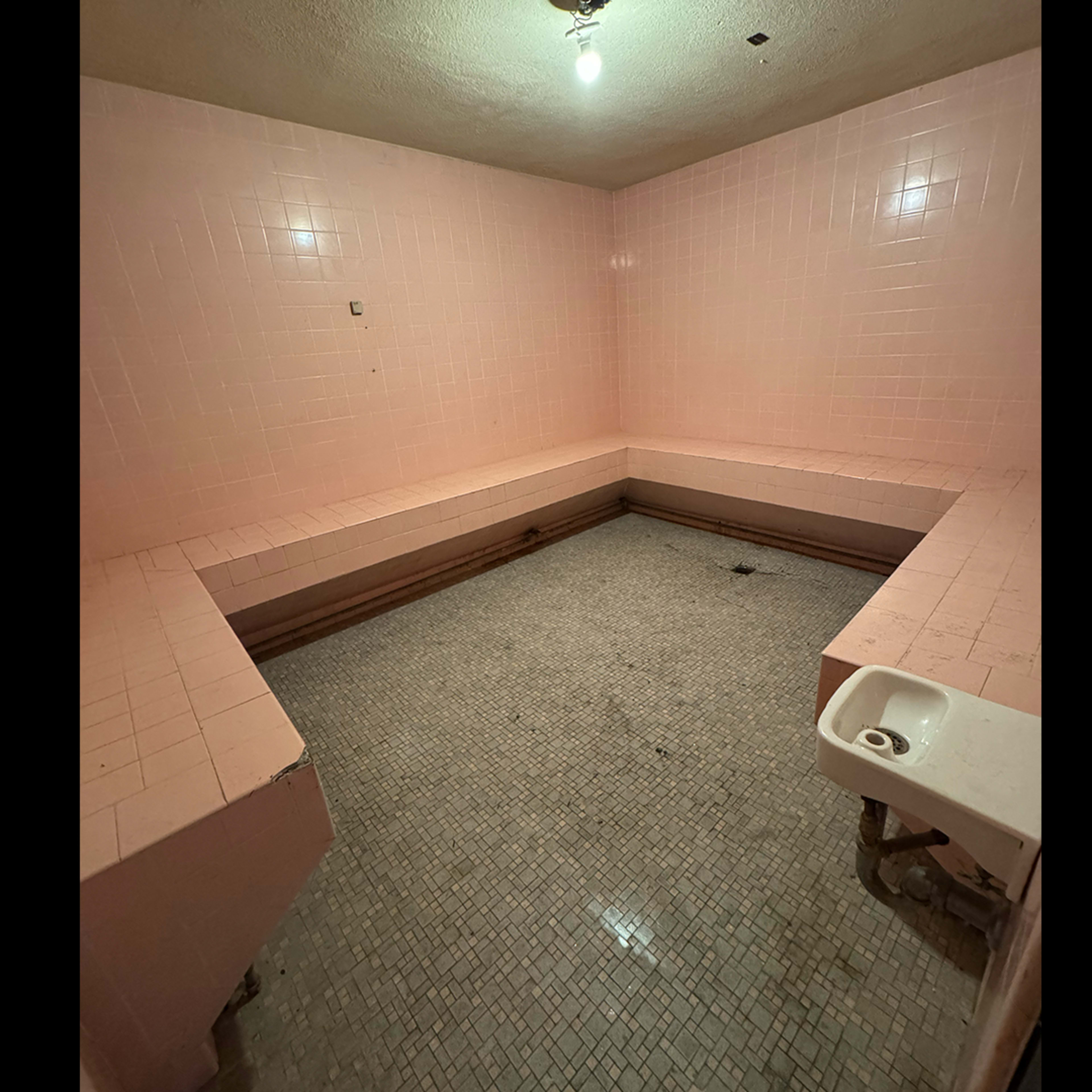 The image shows a vacant, tiled room with pink walls and a sink in one corner, featuring benches along the sides and a tiled floor.