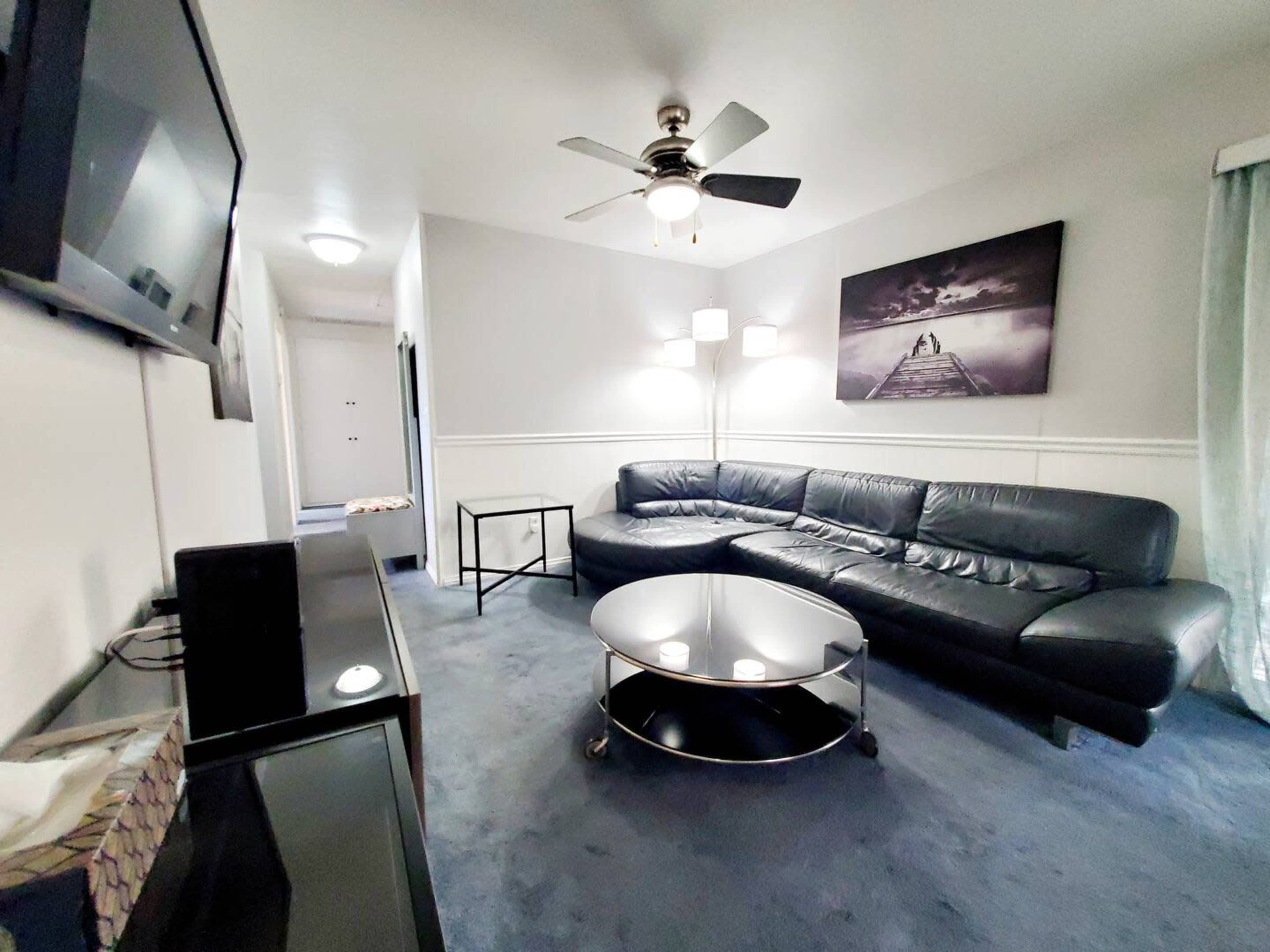 A living room featuring a black leather sectional sofa, a round coffee table, a television on a console, and light fixtures, all set against a light-colored wall.