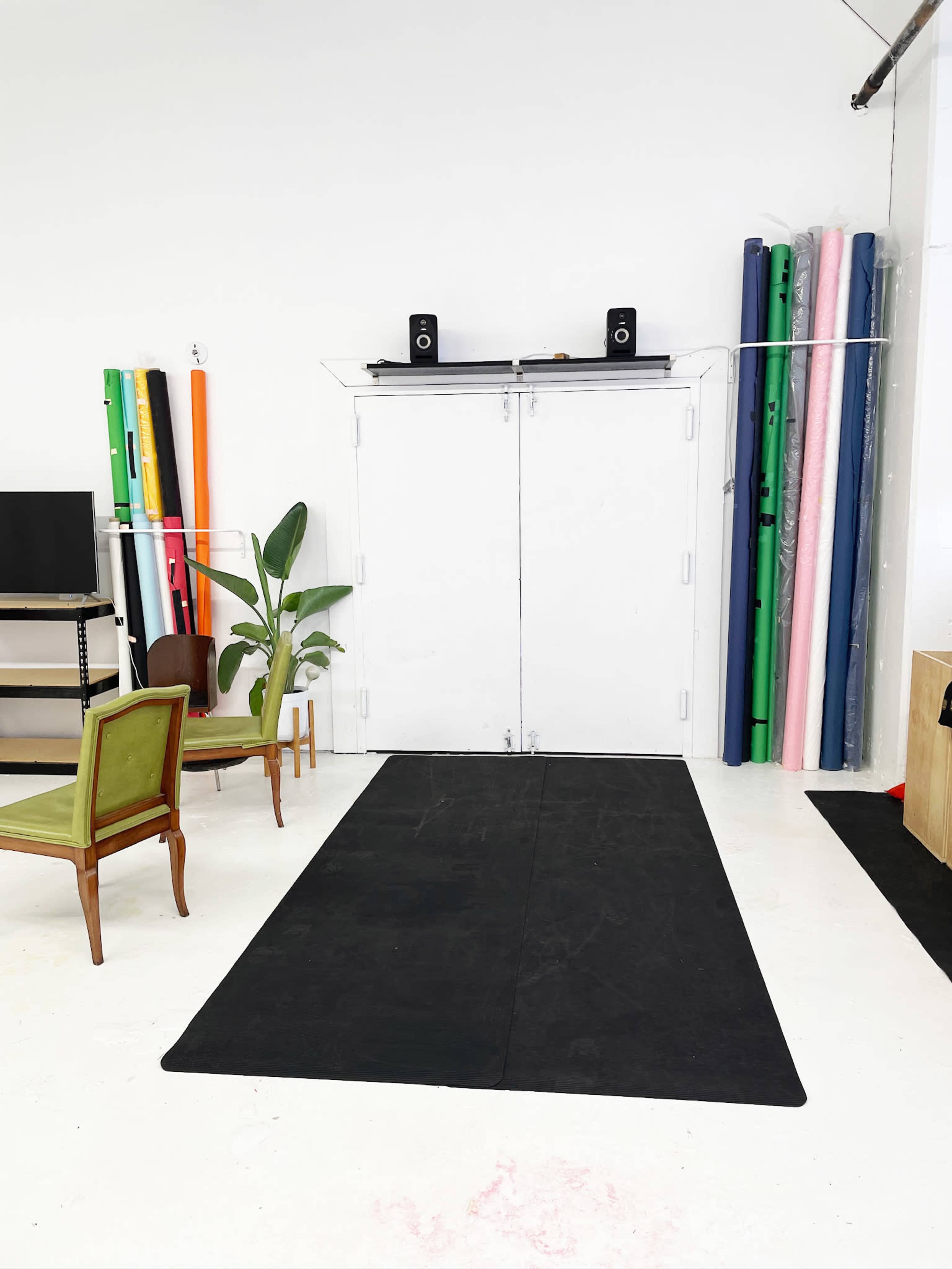 The image shows a bright, minimalist studio space with a black mat on the floor, two green chairs, a television stand, and colorful rolls of material stacked against the wall.