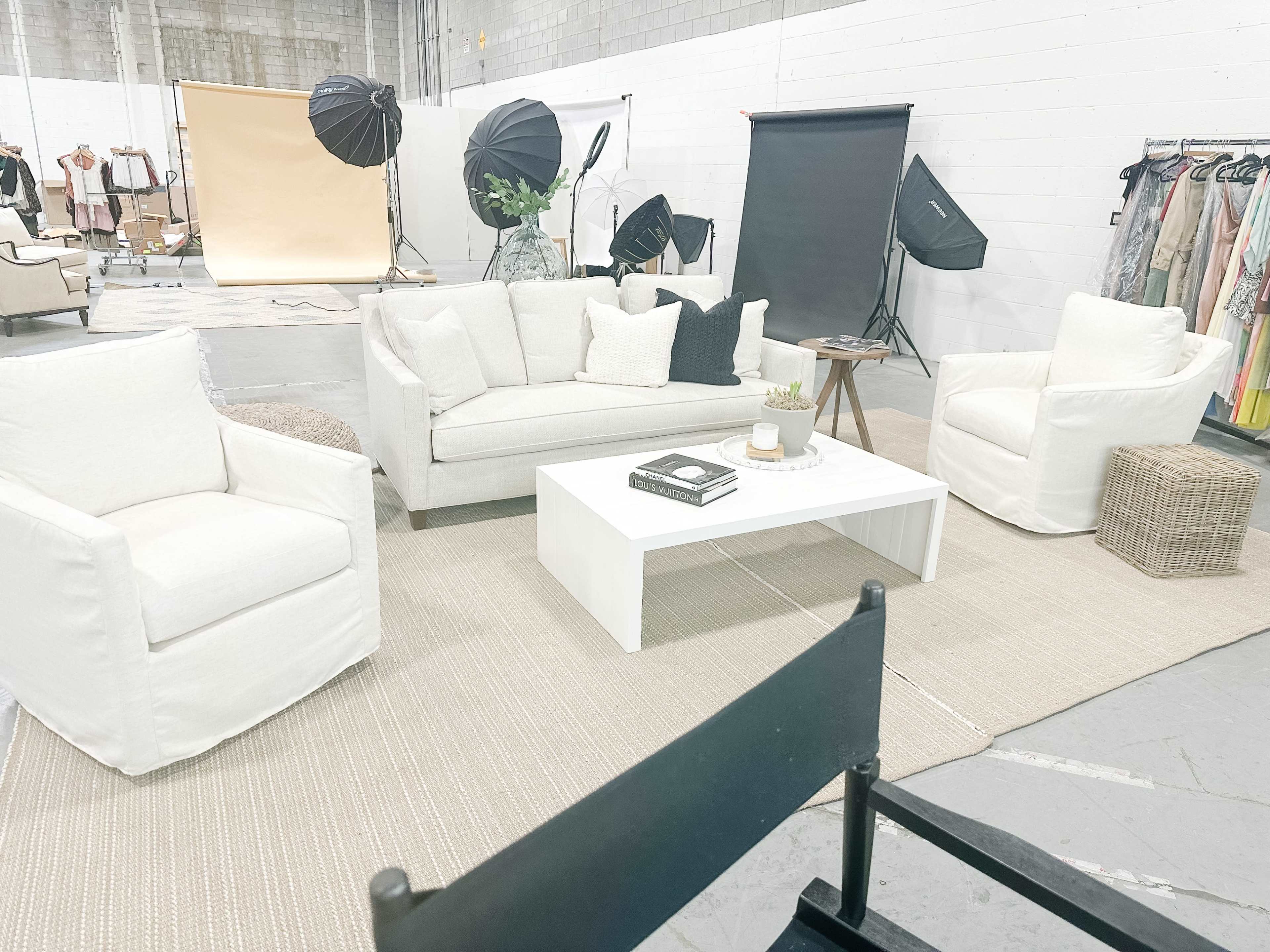 The scene shows a styled photo shoot space with white furniture, a coffee table, and photography equipment set up in a large, industrial-style room.