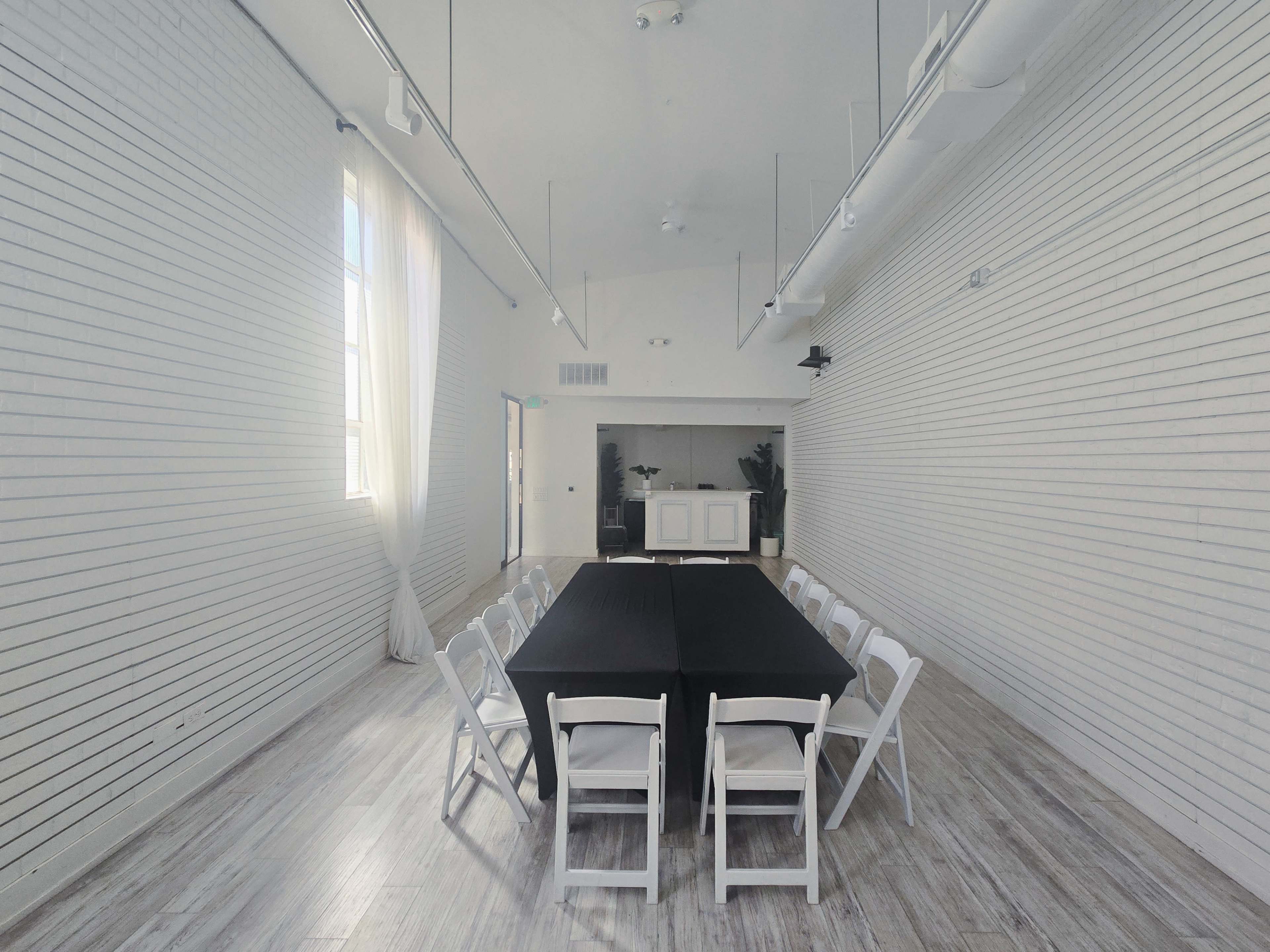A rectangular black table is set with white folding chairs in a brightly lit, minimalist meeting room with white walls and wooden flooring.