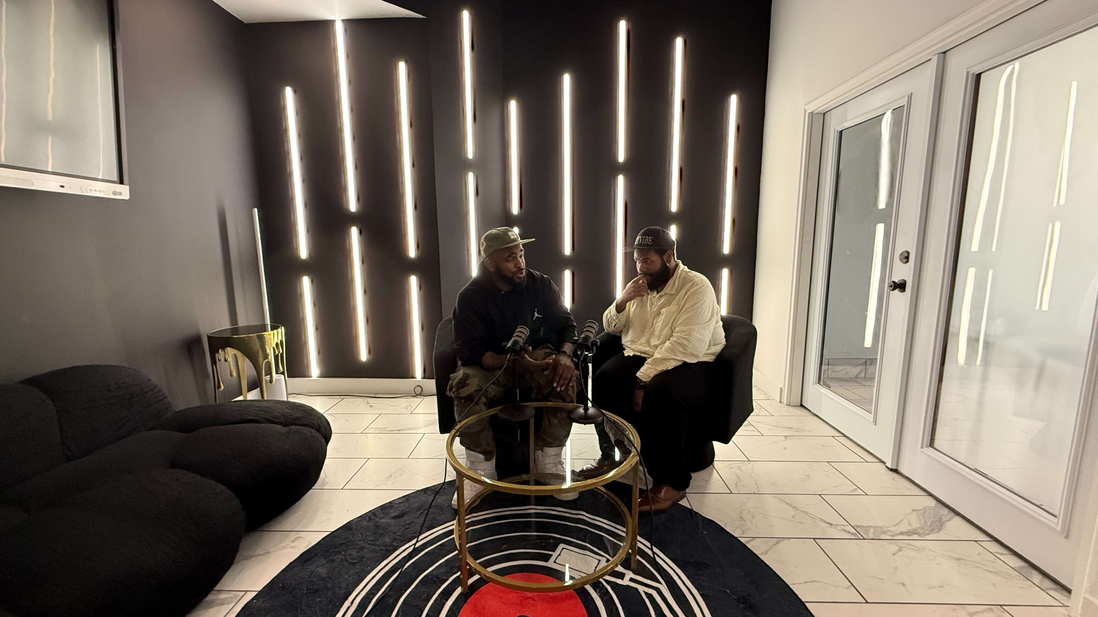 Two men sit near a table with microphones in a modern room featuring a black wall with vertical light strips and a white door.