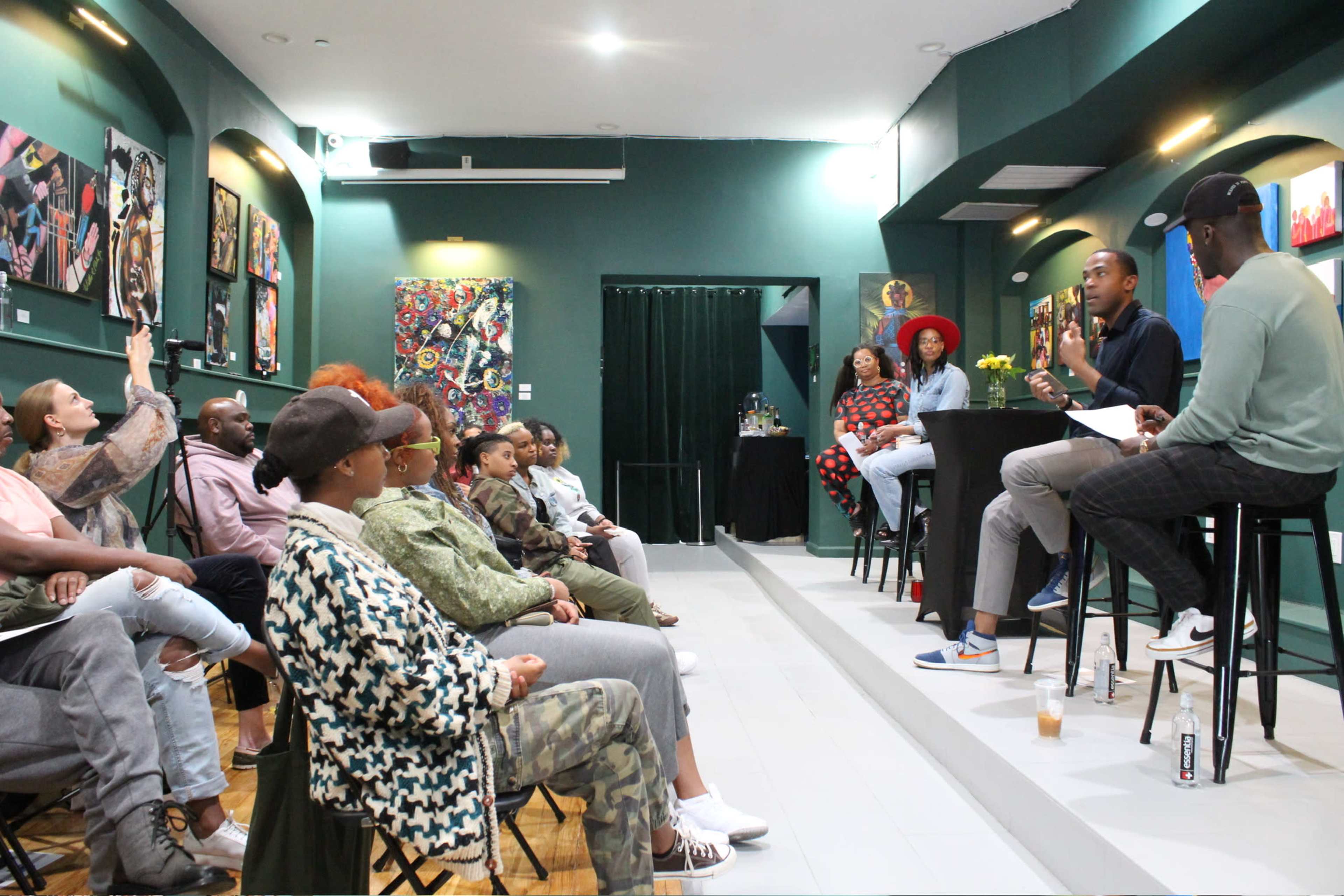 A group of people is seated in a modern venue with colorful artwork on the walls, engaging in a discussion while a speaker presents at the front.