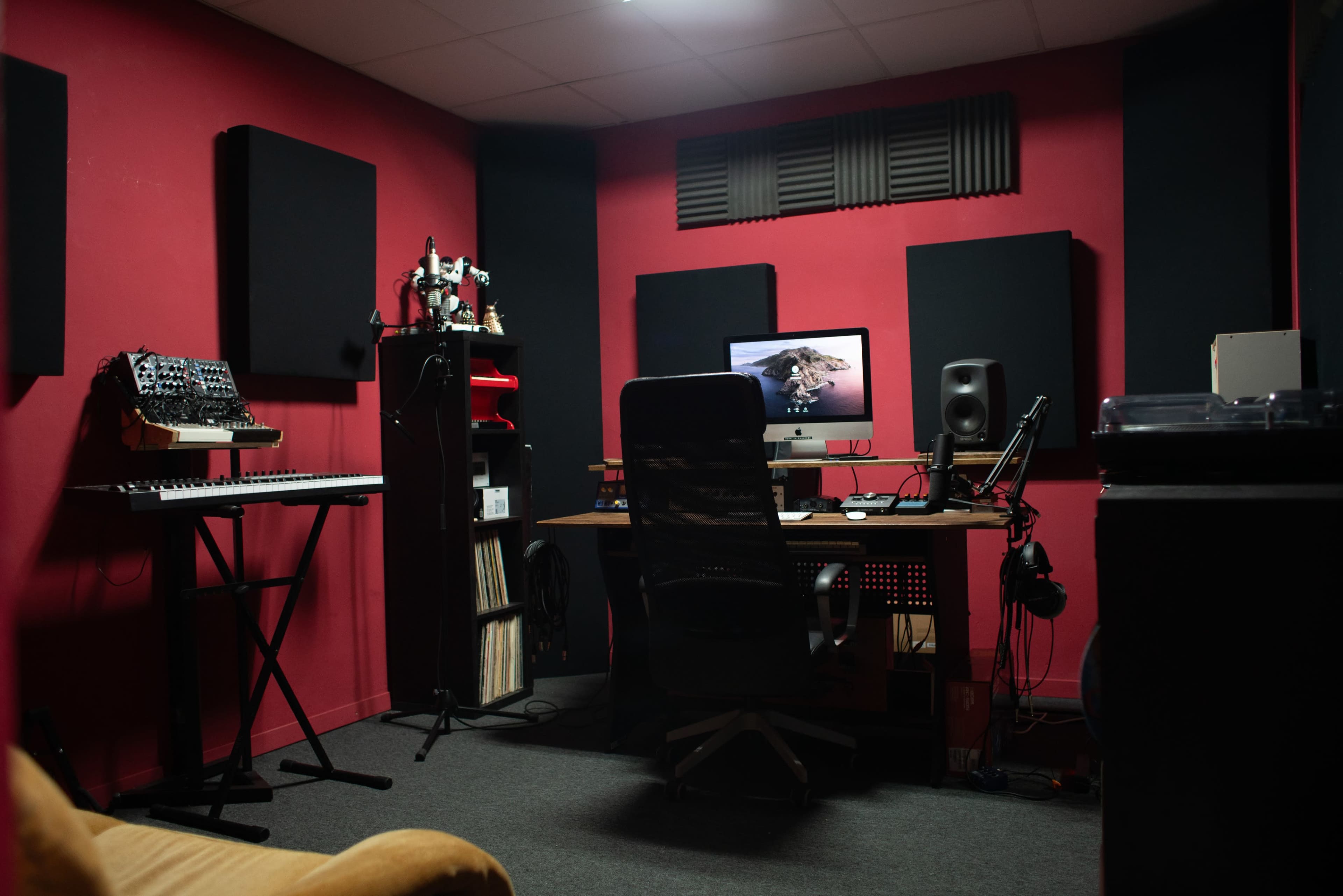 A music production room features a keyboard, an audio workstation with a monitor, soundproofing panels, and various recording equipment.