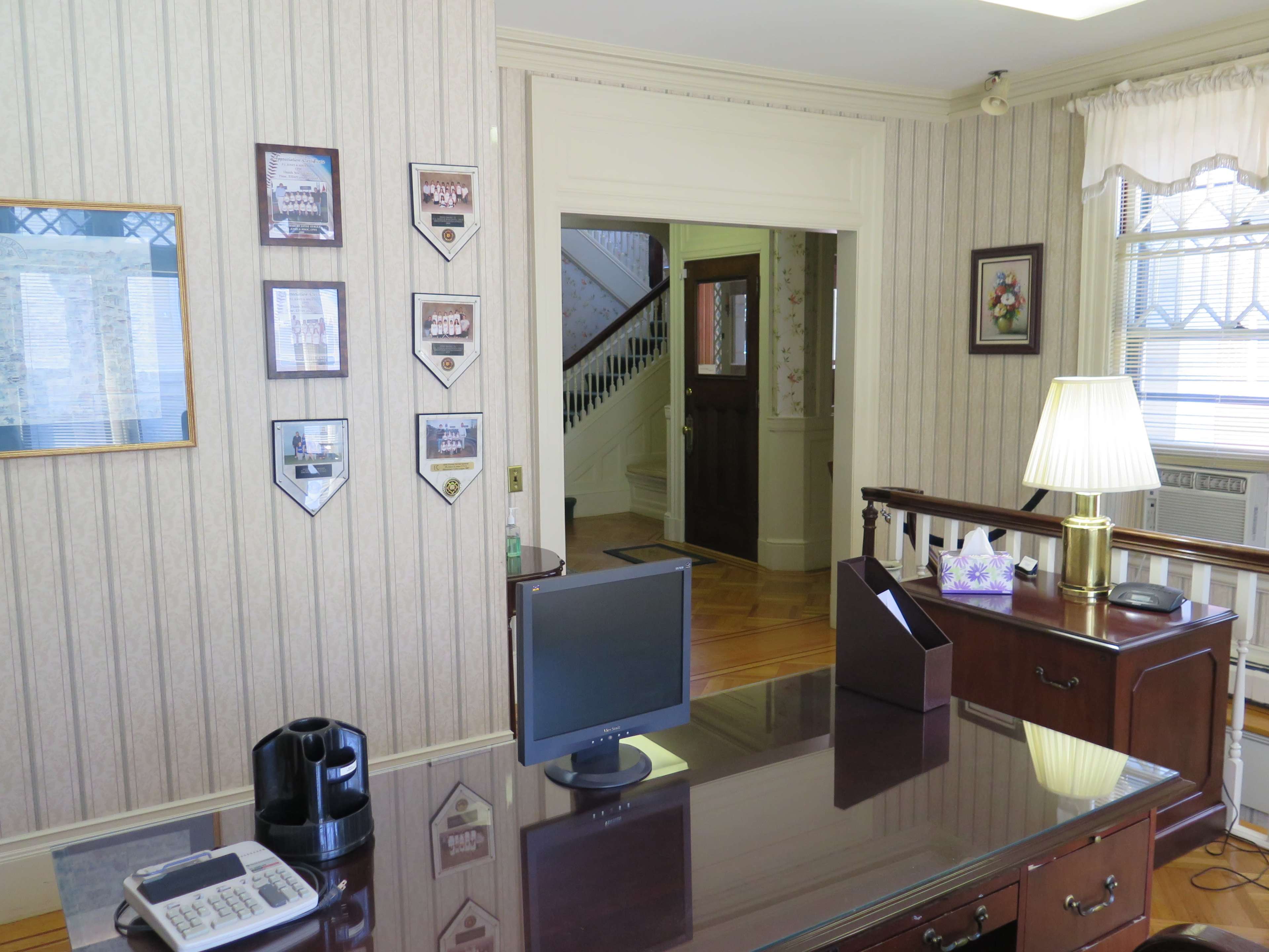 The image shows an office with a wooden desk, a computer monitor, a lamp, and framed wall decorations, leading to a doorway with stairs visible in the background.