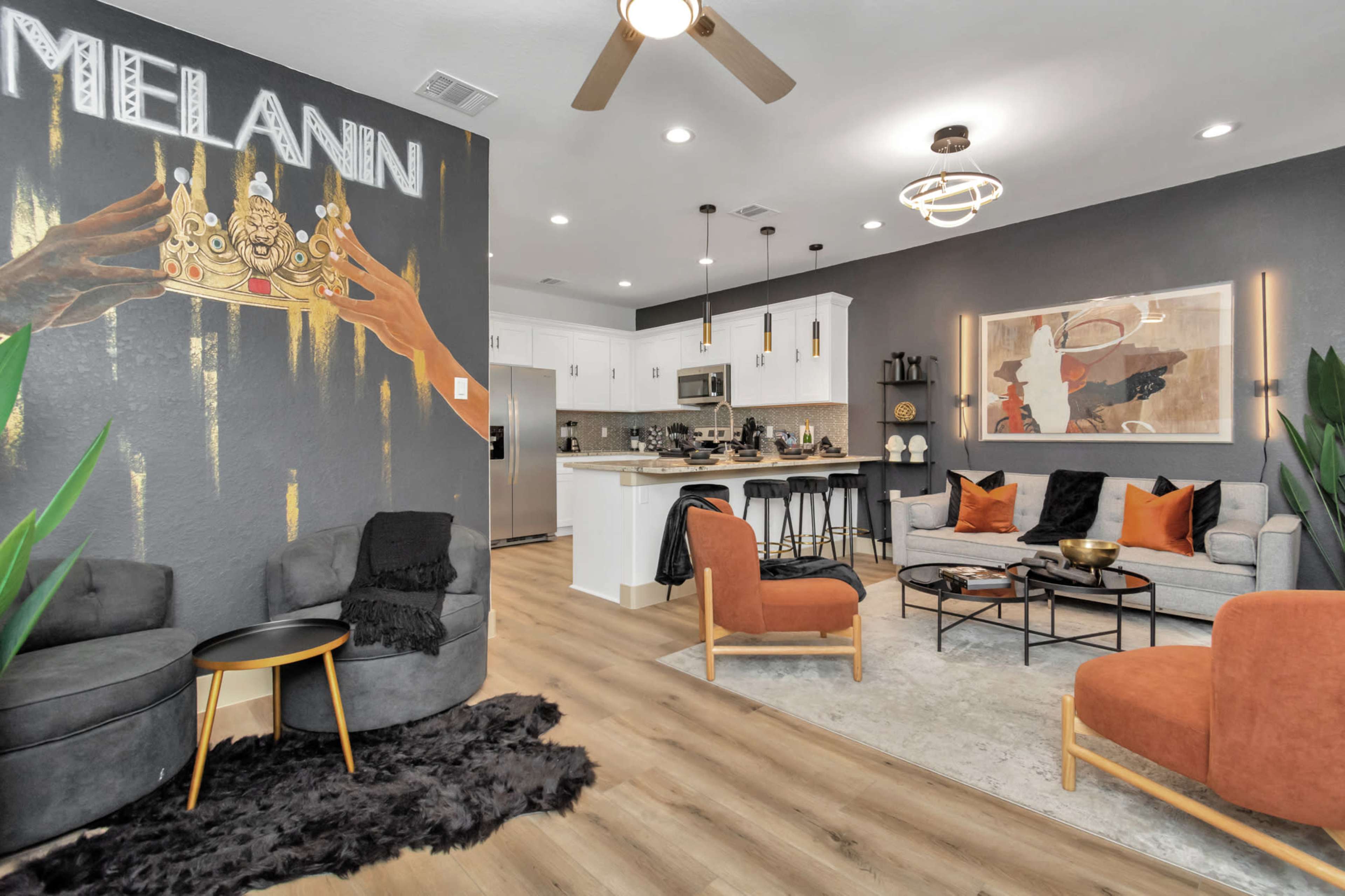 The image shows a stylish living area with modern furniture, a kitchen visible in the background, and a wall featuring artwork that highlights the word "MELANIN."