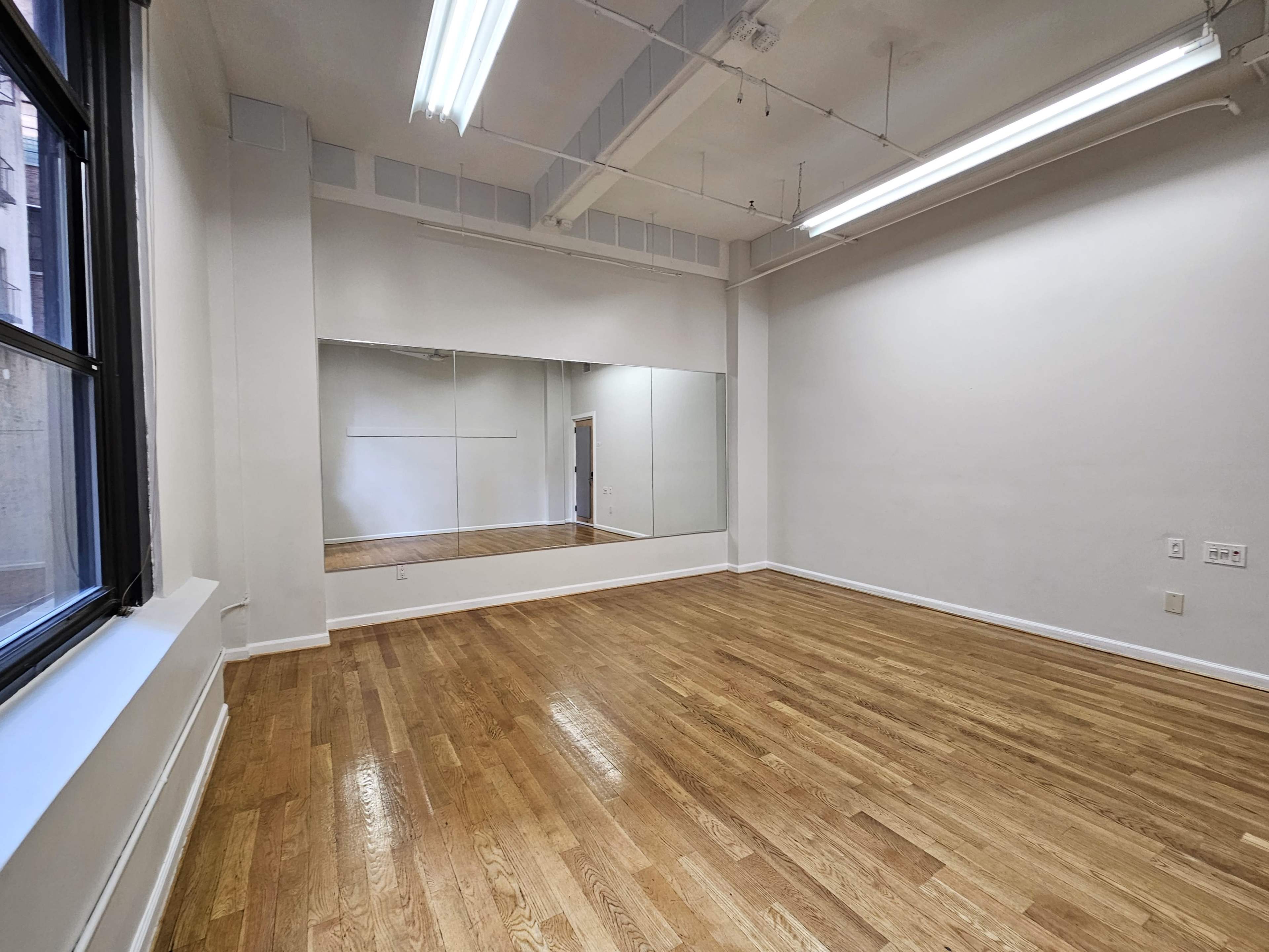 An empty room with hardwood floors and a large wall mirror, illuminated by fluorescent lights.