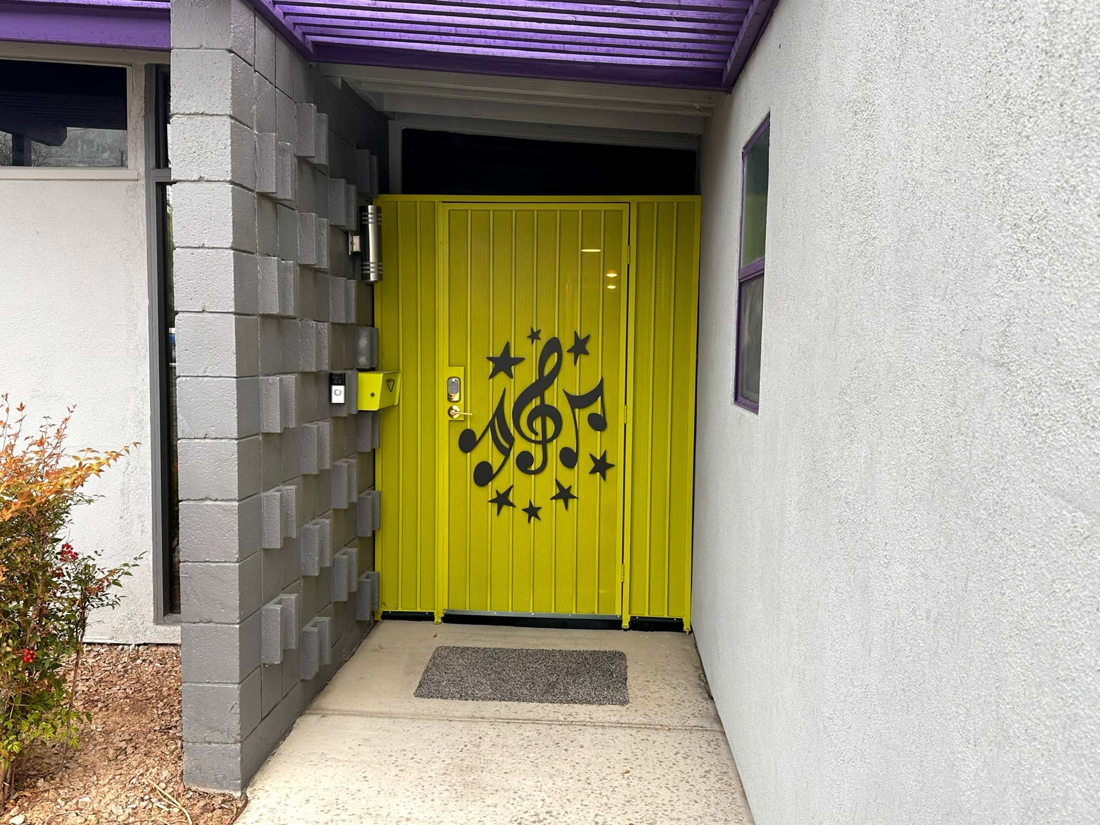 A bright yellow door with musical note designs leads into a gray building, framed by a purple overhang and a concrete block wall.