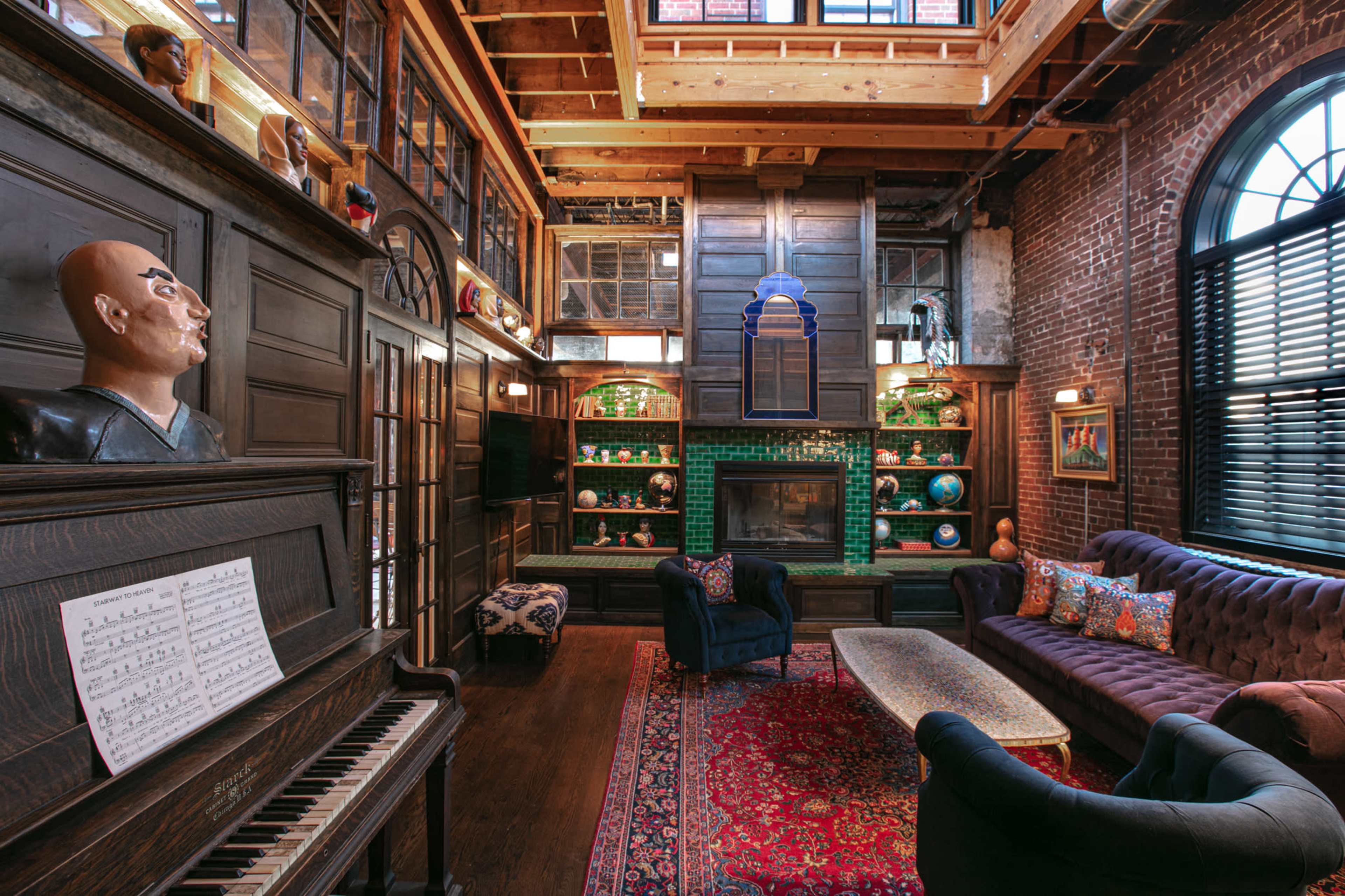 The image depicts a spacious living room with exposed brick walls, wooden beams, and a grand piano, featuring dark blue armchairs, a plush sofa, and shelves adorned with decorative items.