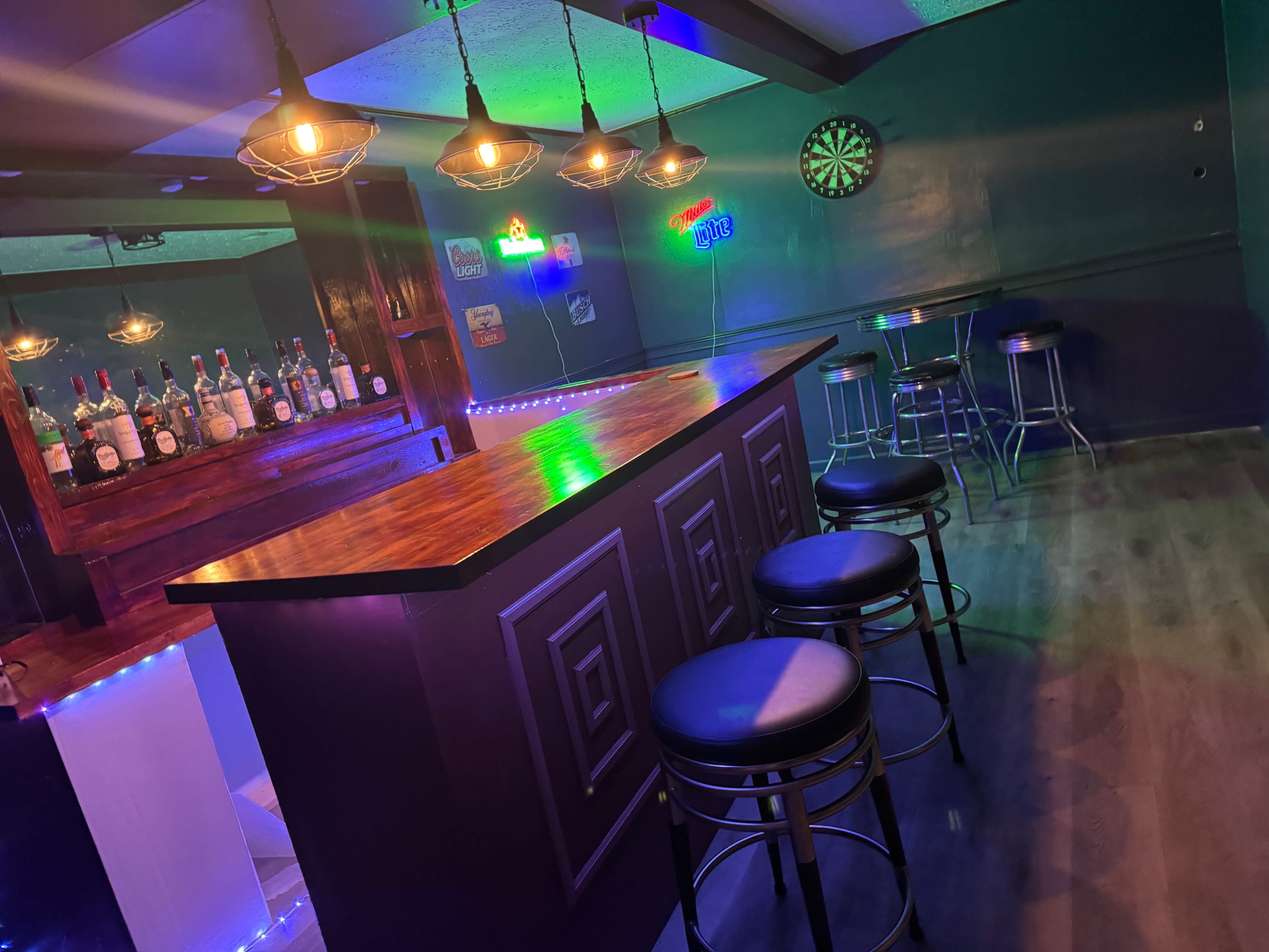 A home bar features a wooden counter with four black stools, dim lighting, and shelves stocked with various bottles in the background.