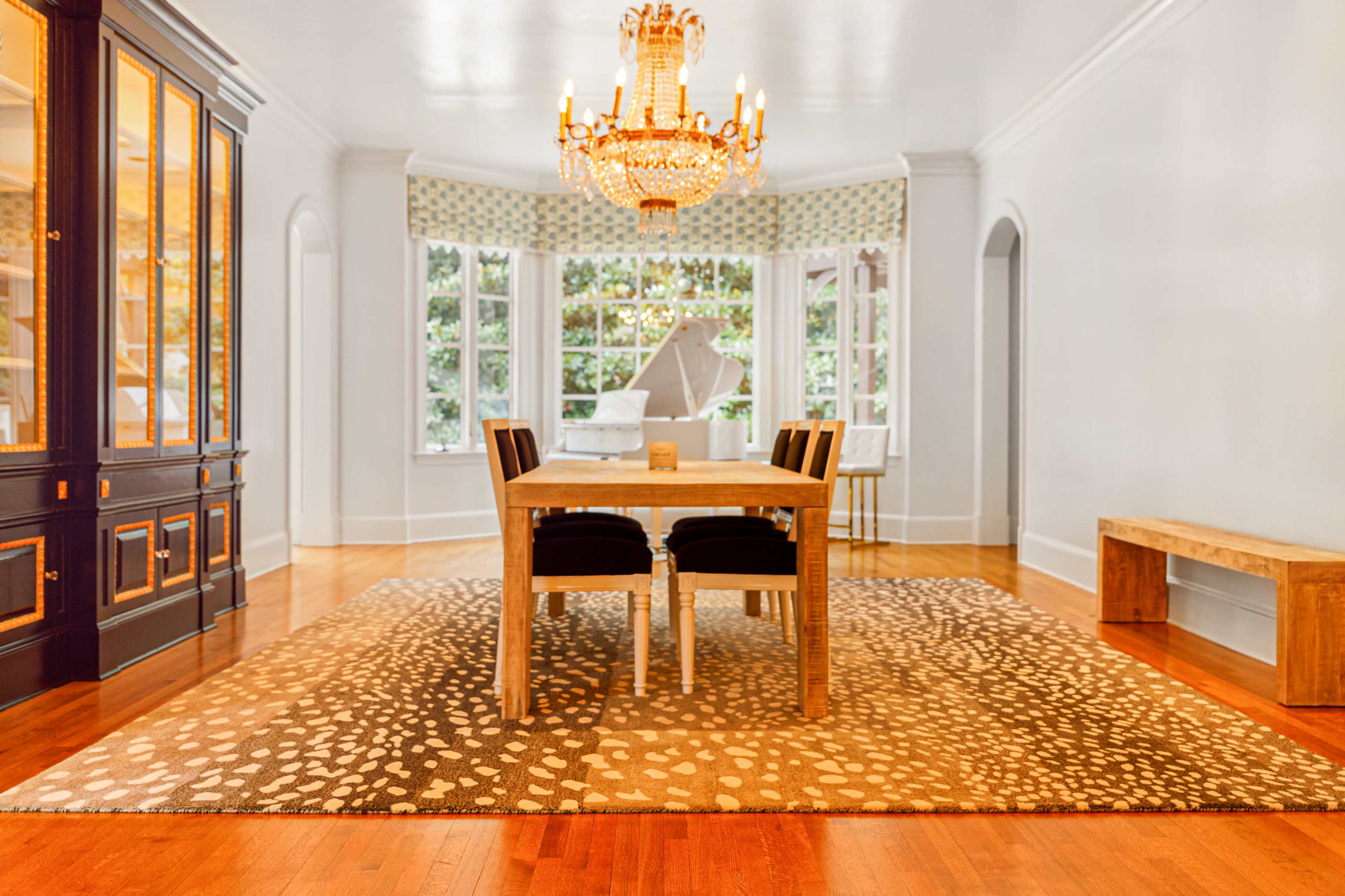 A dining area features a wooden table surrounded by black chairs, set on a patterned rug, with a chandelier overhead and large windows letting in natural light.