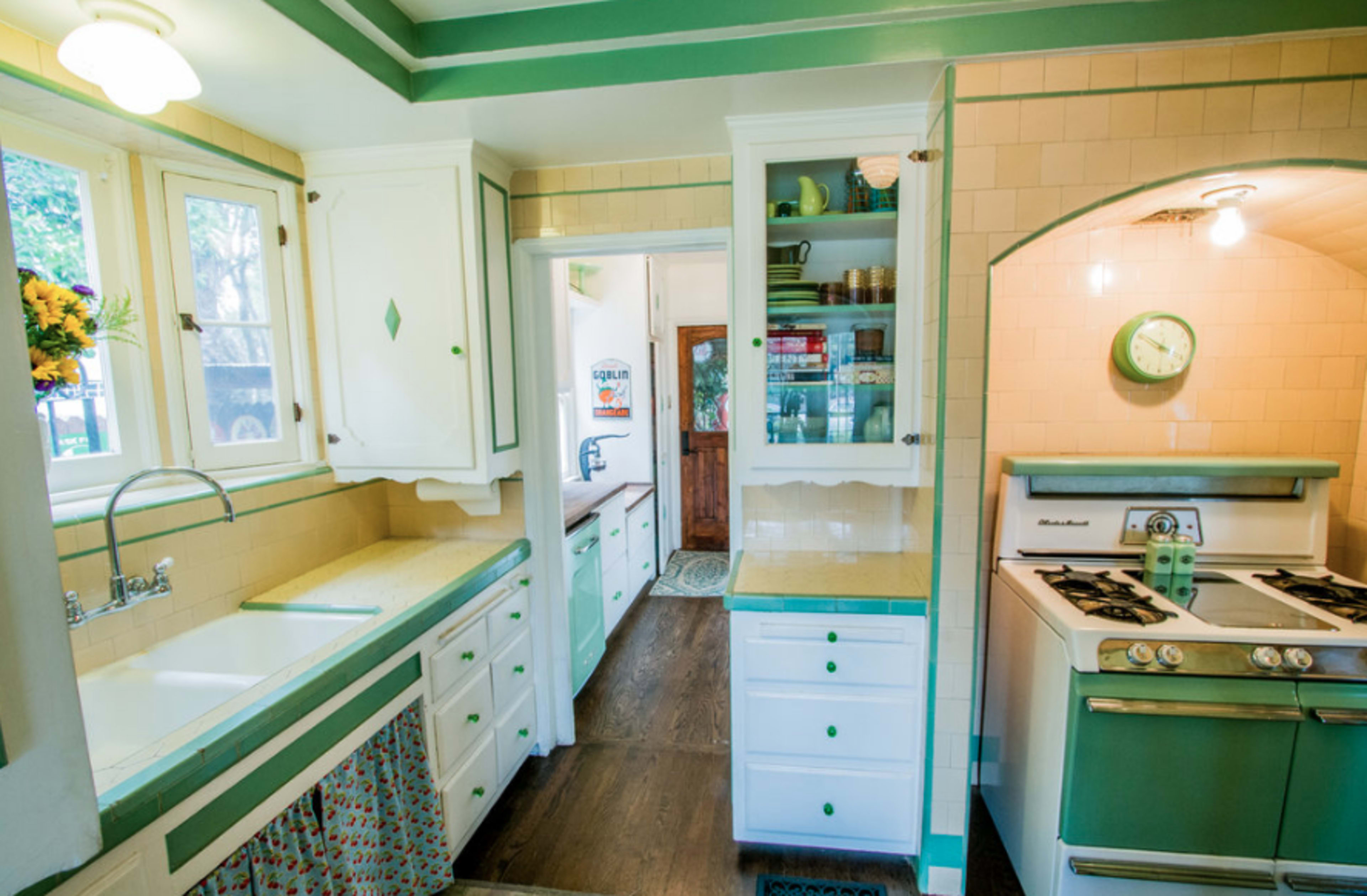 A vintage kitchen with white and green cabinetry, a double sink, and a gas stove, all arranged in a light-filled space with tiled walls.