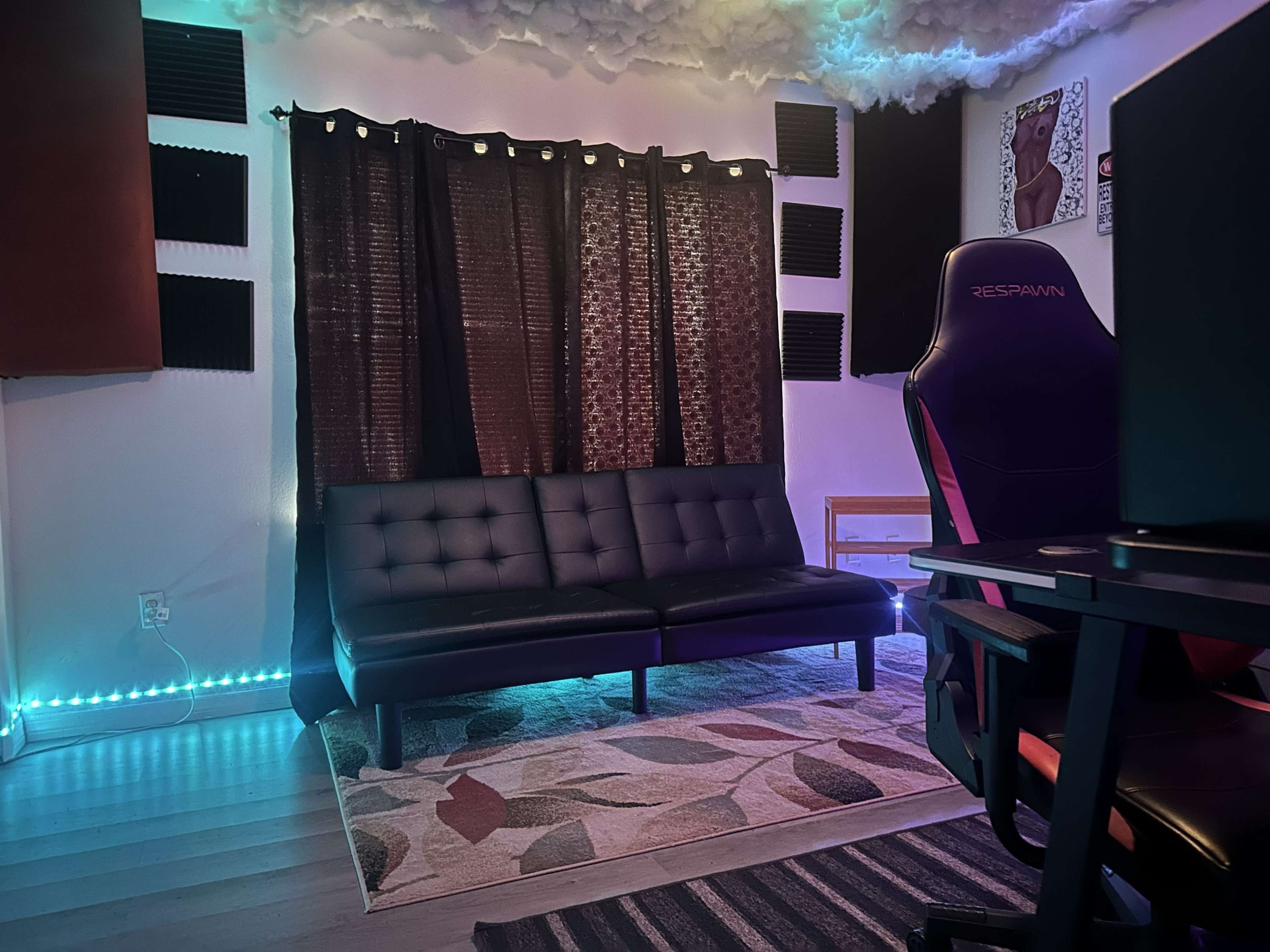The image shows a modern room with a black sofa, soft lighting, a patterned rug, and a workspace featuring a gaming chair and a computer desk.