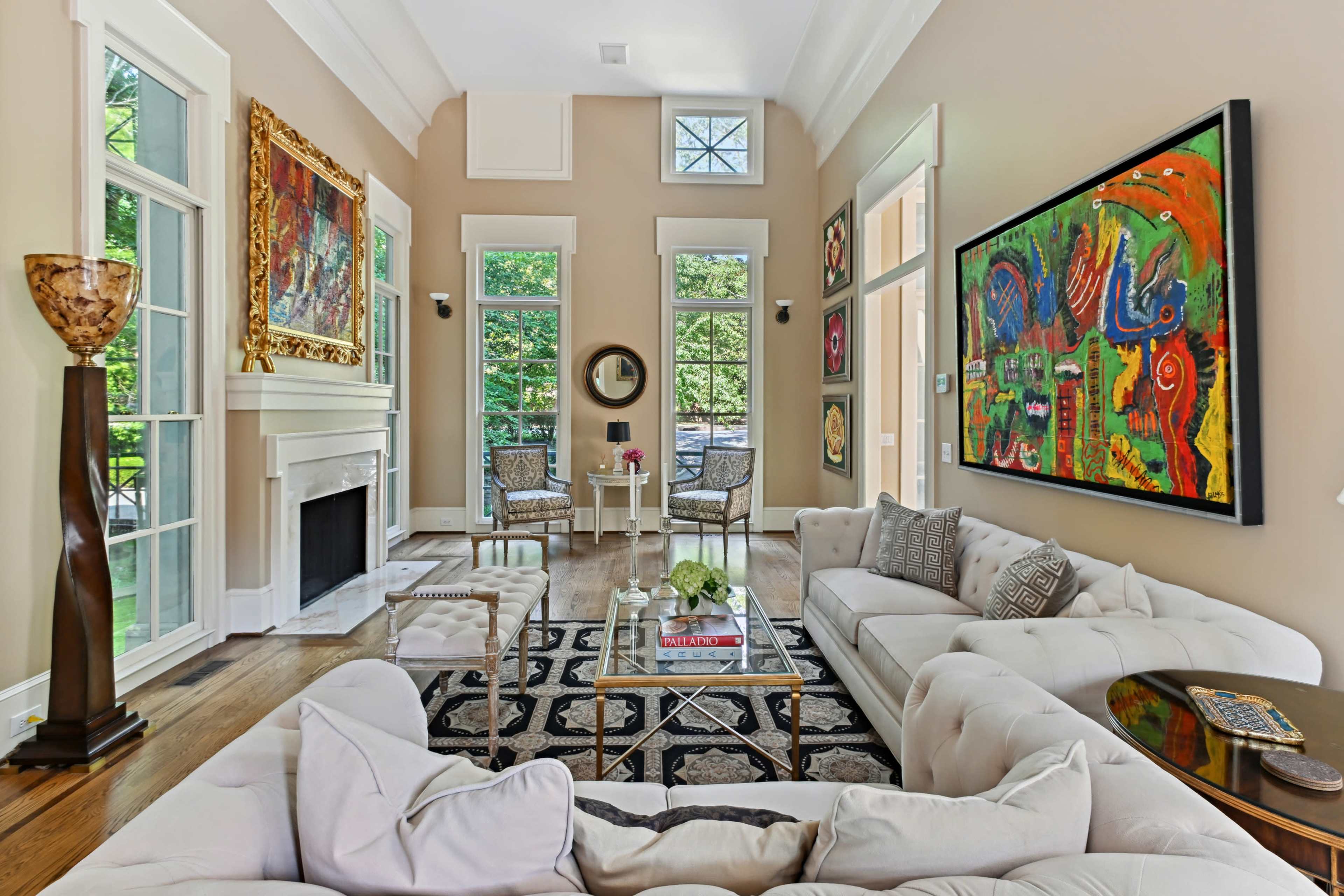 A well-lit living room featuring a large artwork on the wall, a white sofa, a coffee table in the center, and large windows overlooking greenery.