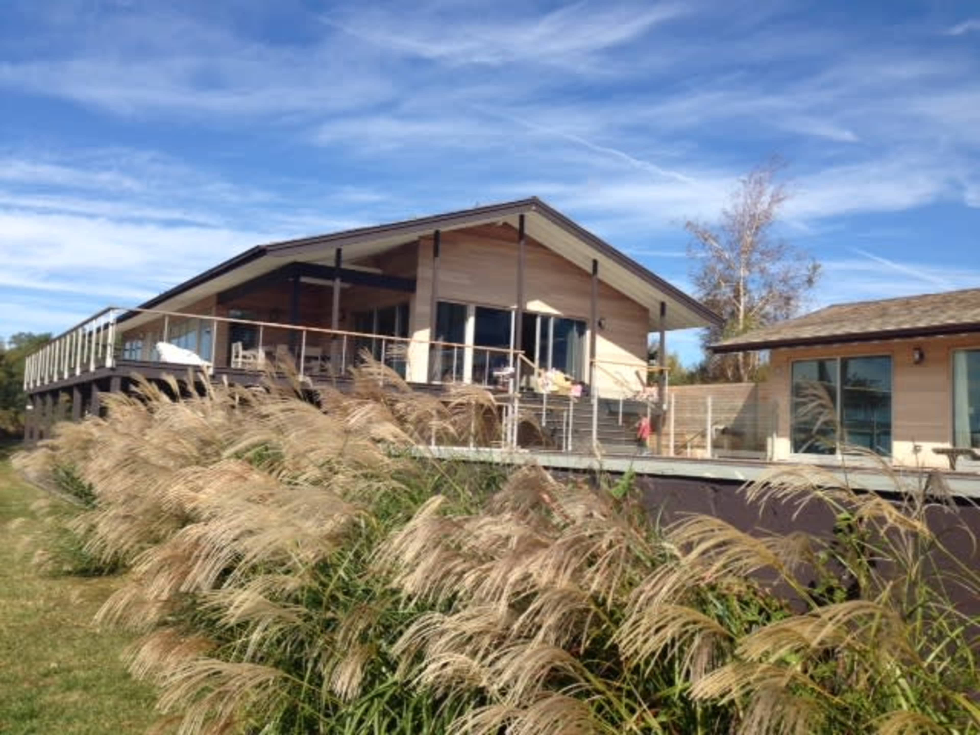 A modern house with a large deck is situated beside tall grass under a clear blue sky.