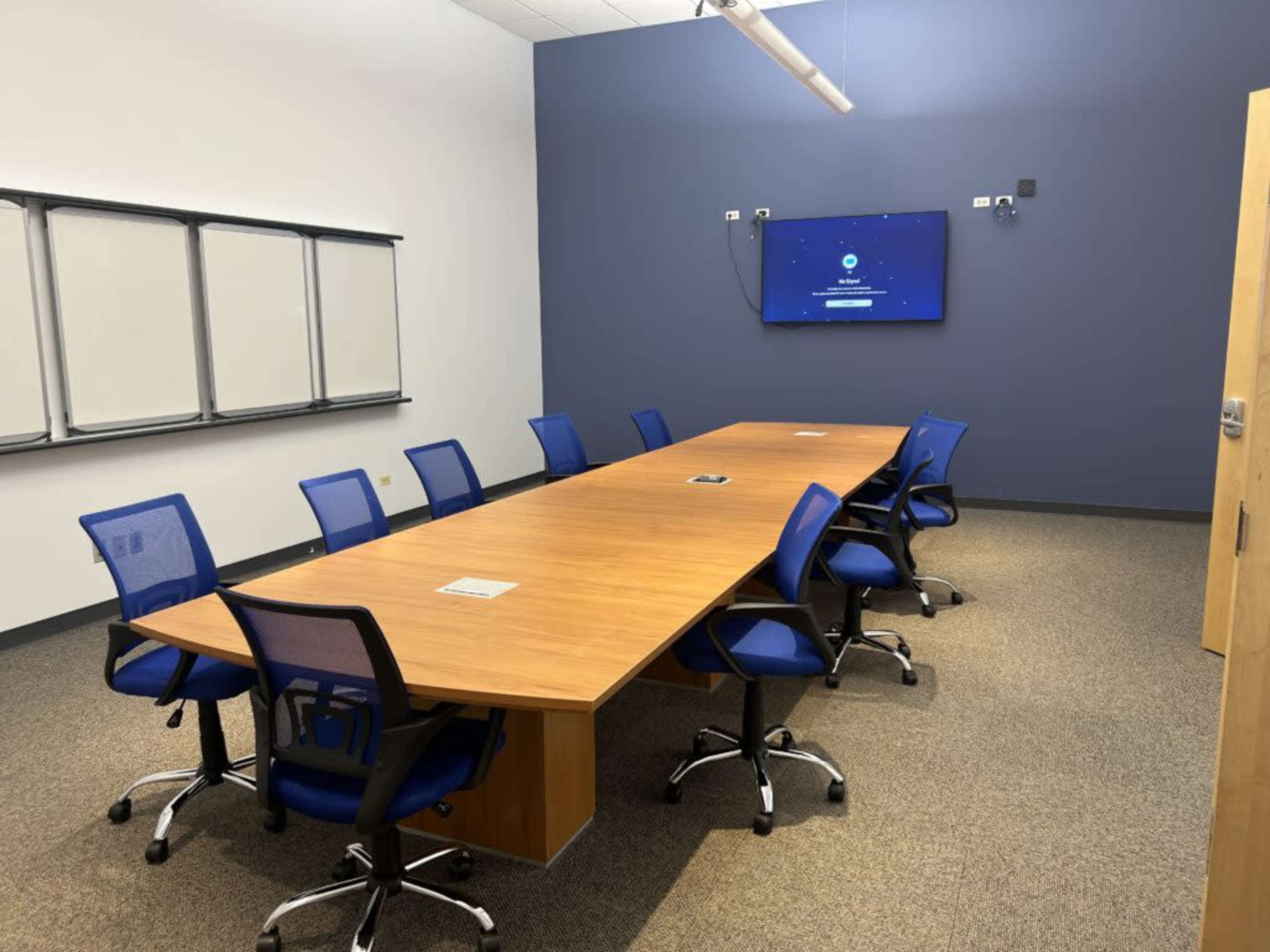 The image shows a modern conference room with a long wooden table, blue chairs, and a wall-mounted screen displaying a welcome message.