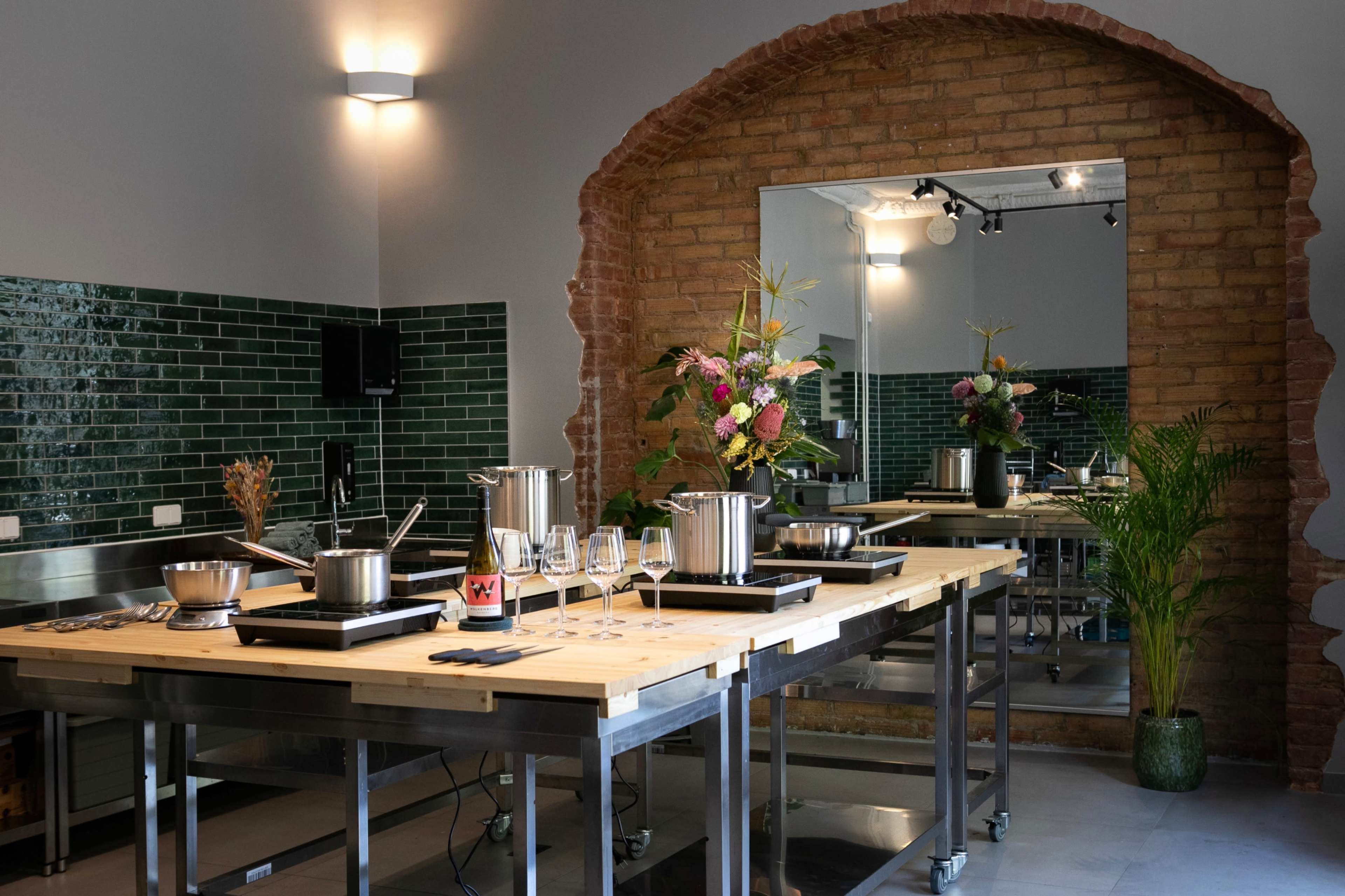 The image shows a modern cooking classroom with stainless steel tables, pots on induction cooktops, and a large mirror framed by a brick arch.