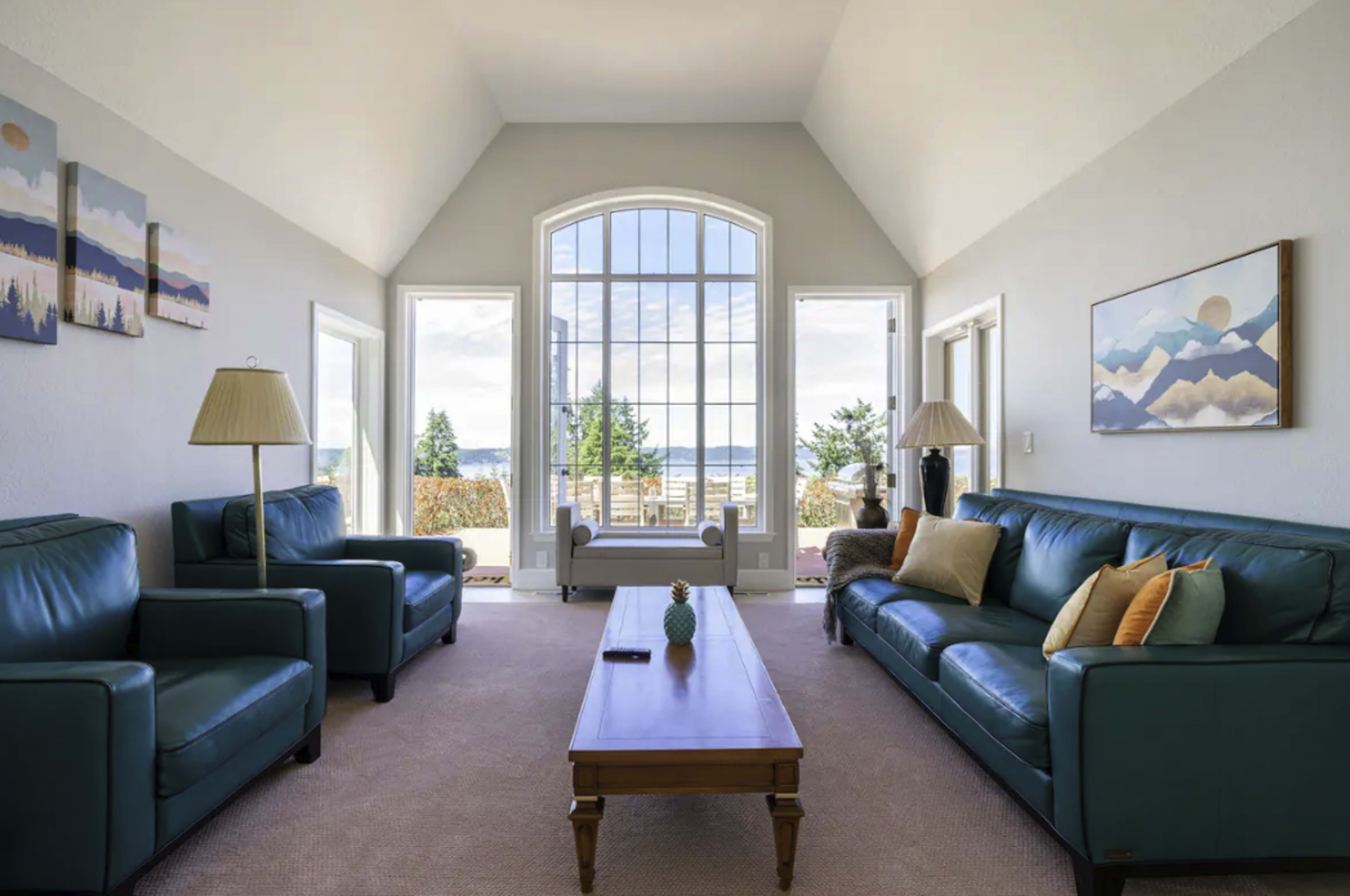 The living room features two teal sofas facing a wooden coffee table, with large windows offering a view of the outdoors.
