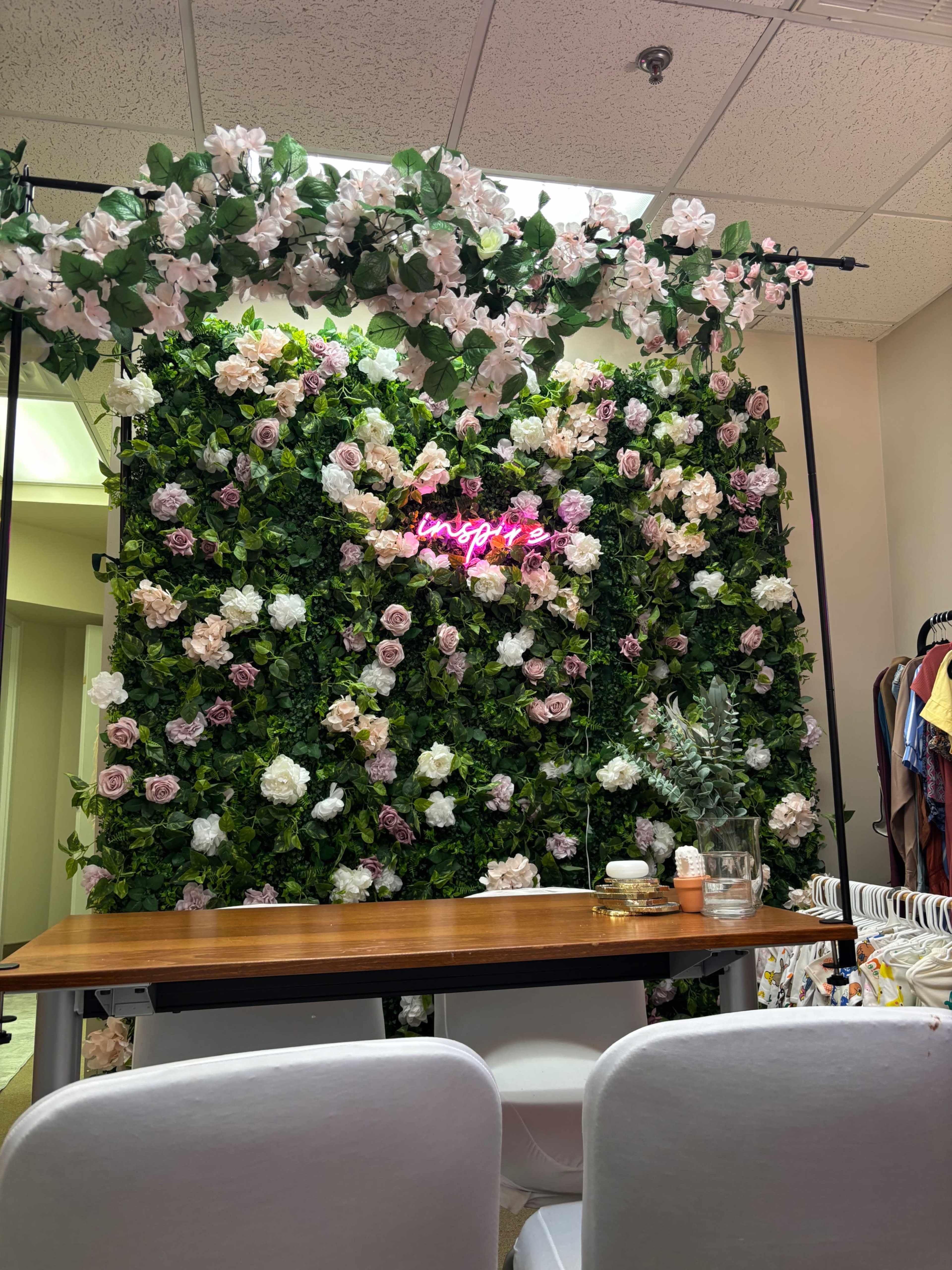 The image shows a floral wall covered in green foliage and pink and white flowers, with a neon sign that reads "insta" glowing in front of a wooden table.