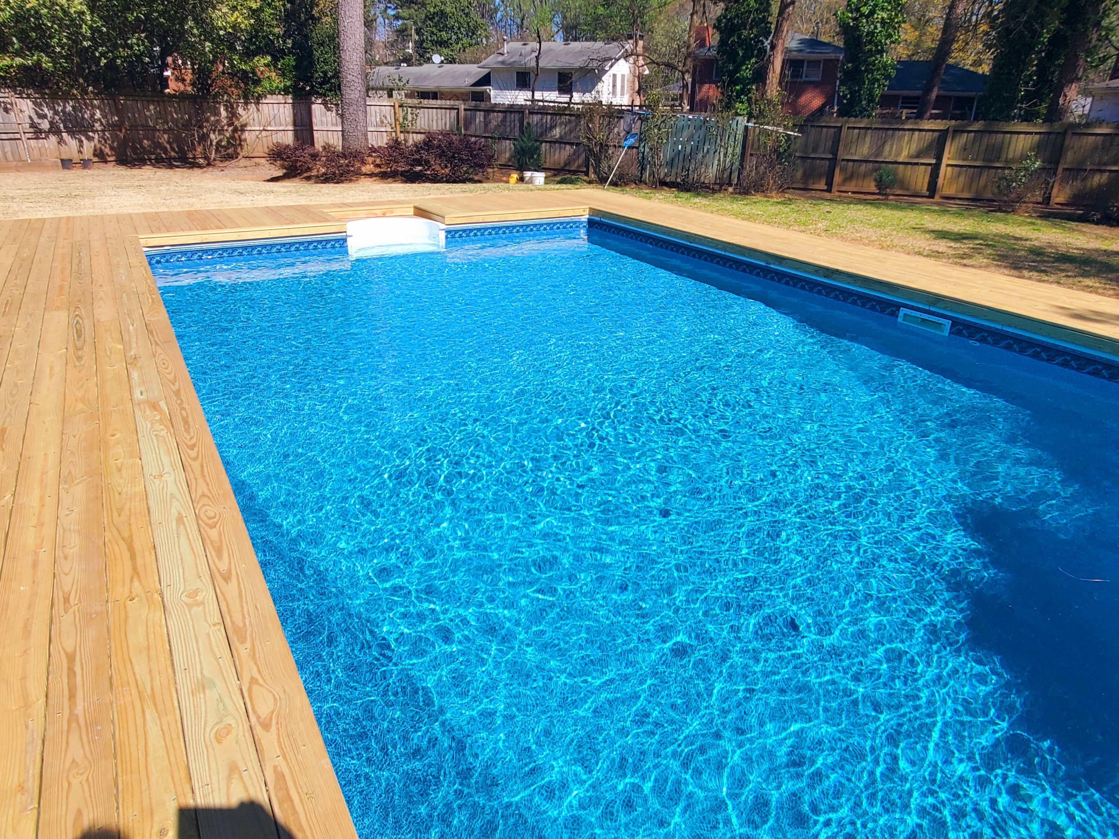 The image shows a clear swimming pool surrounded by wooden decking and a grassy area, with houses and trees in the background.