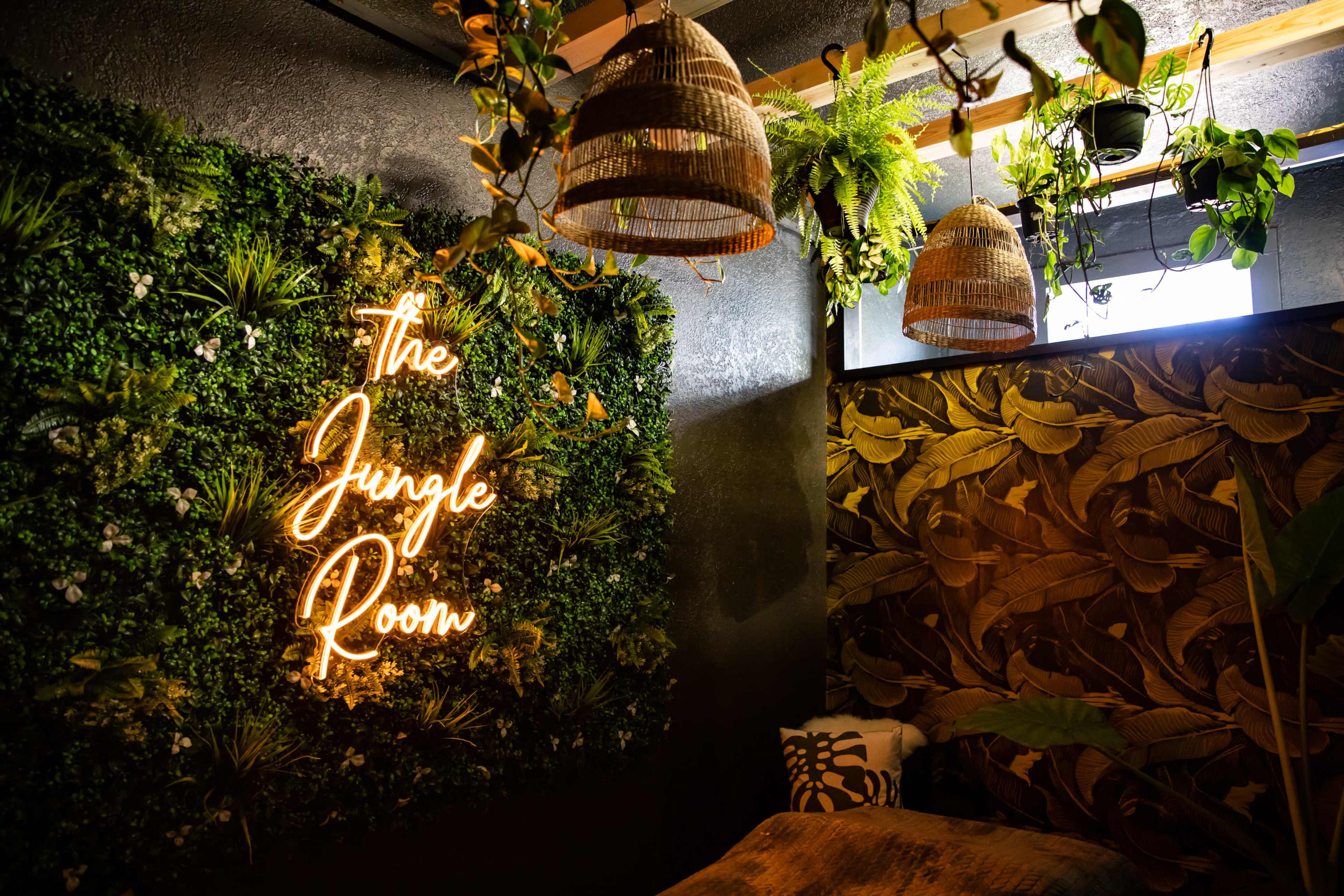 The image shows a room decorated with a neon sign that reads "The Jungle Room," surrounded by greenery and hanging light fixtures.