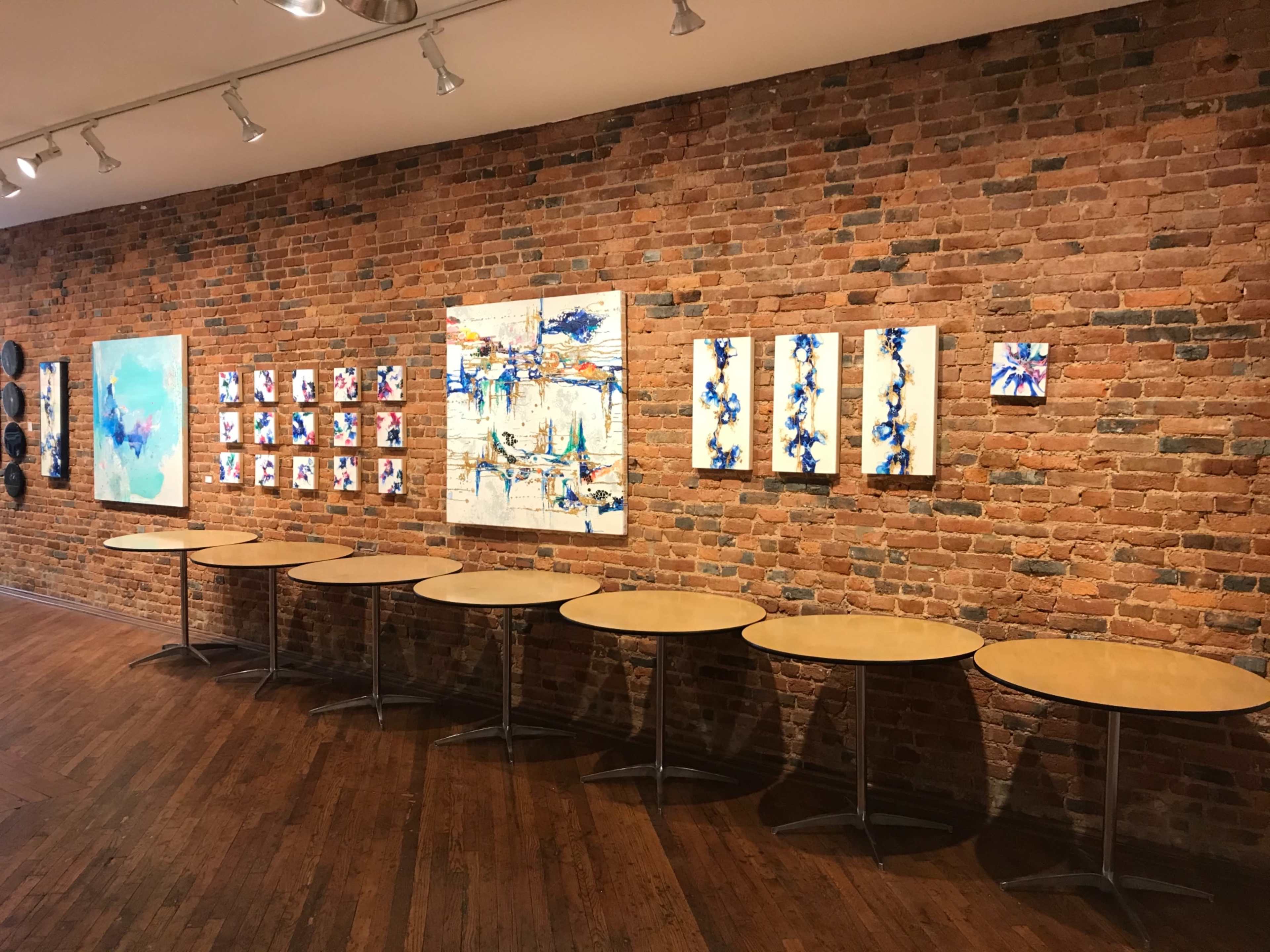 A gallery interior with a brick wall, featuring a series of colorful artworks and several round tables arranged in front.