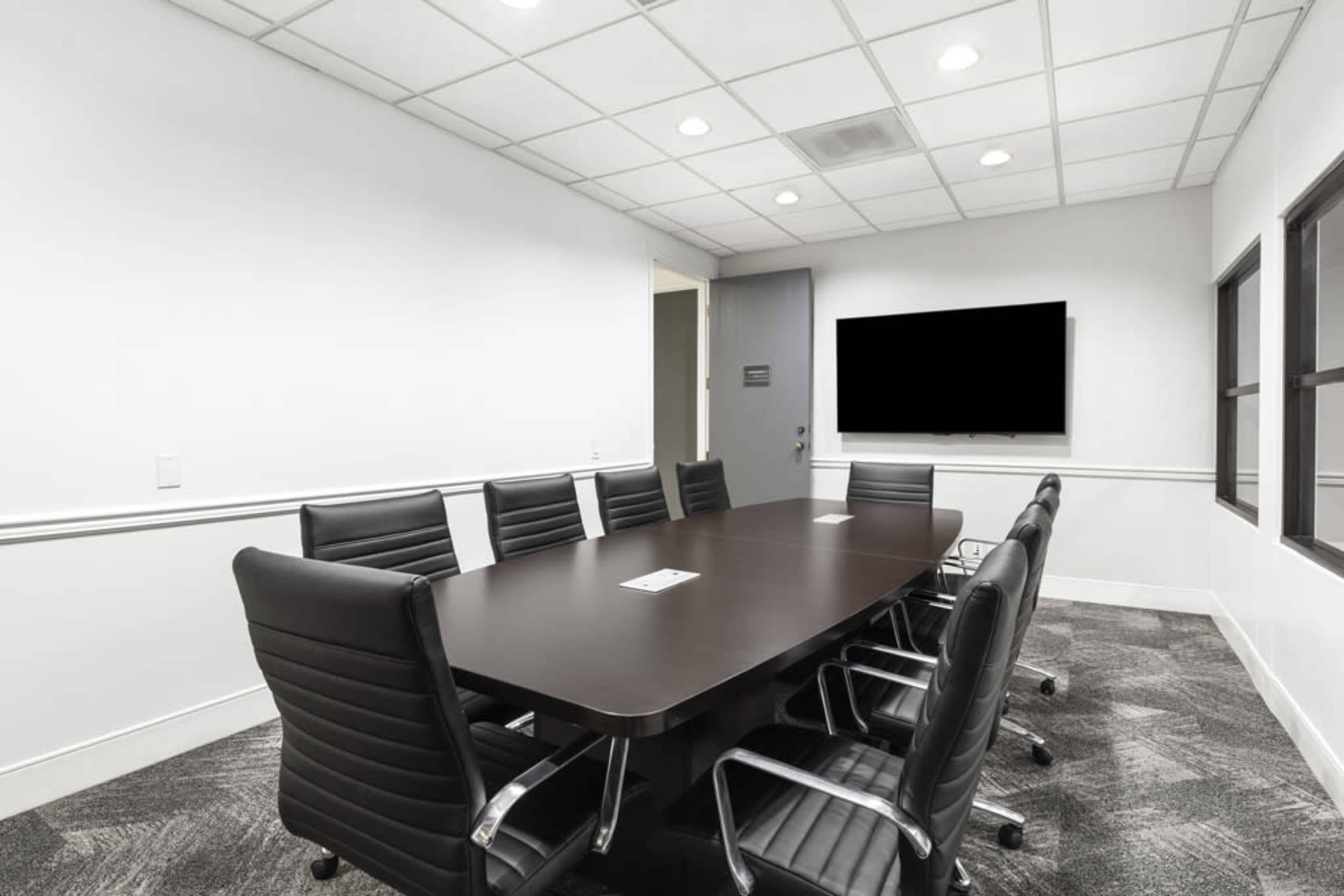 A modern conference room features a large table surrounded by black chairs, with a mounted screen on the wall and windows allowing natural light.