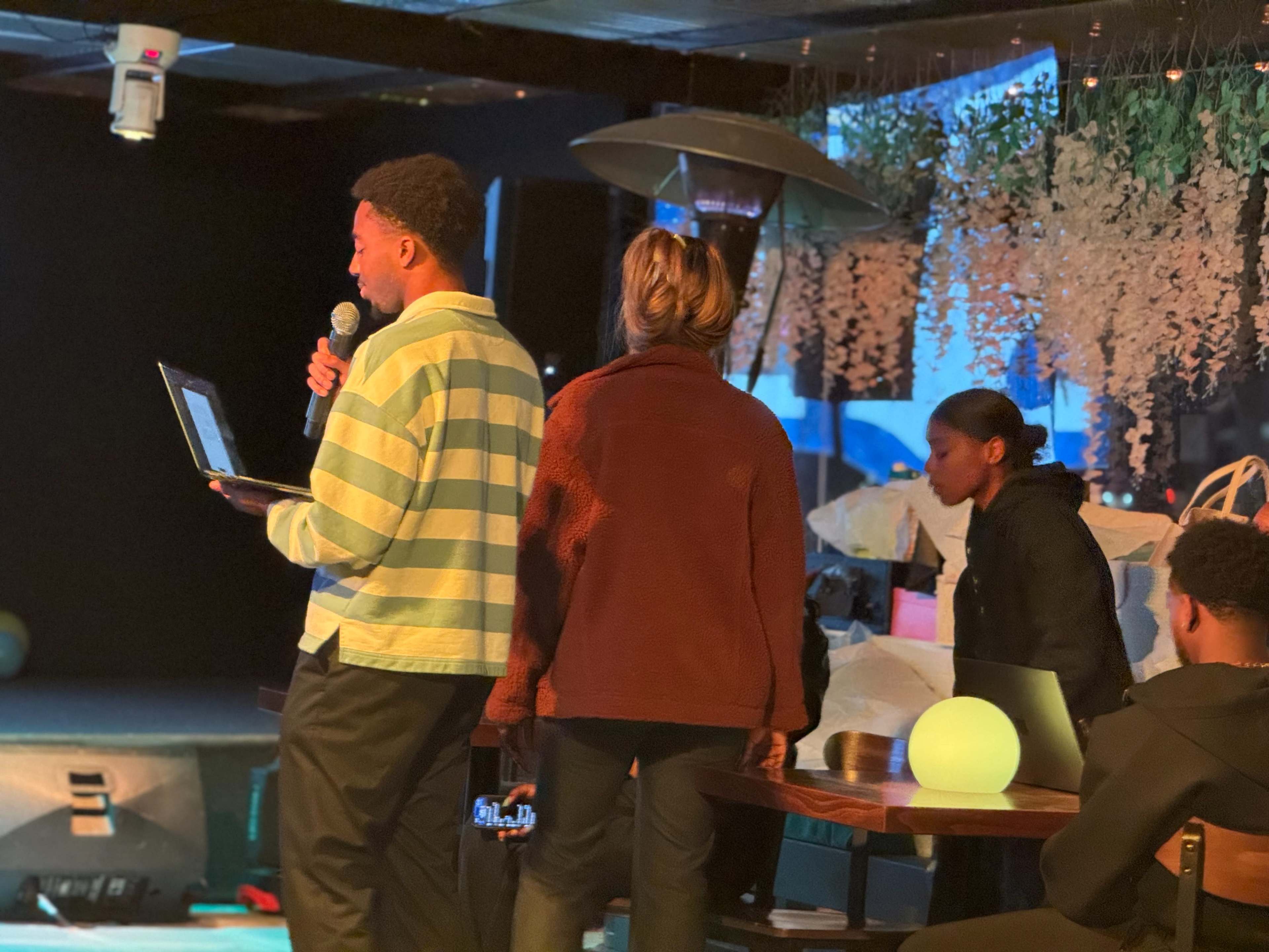 A person holding a microphone stands in front of two others at a small stage, with a laptop open in front of them.