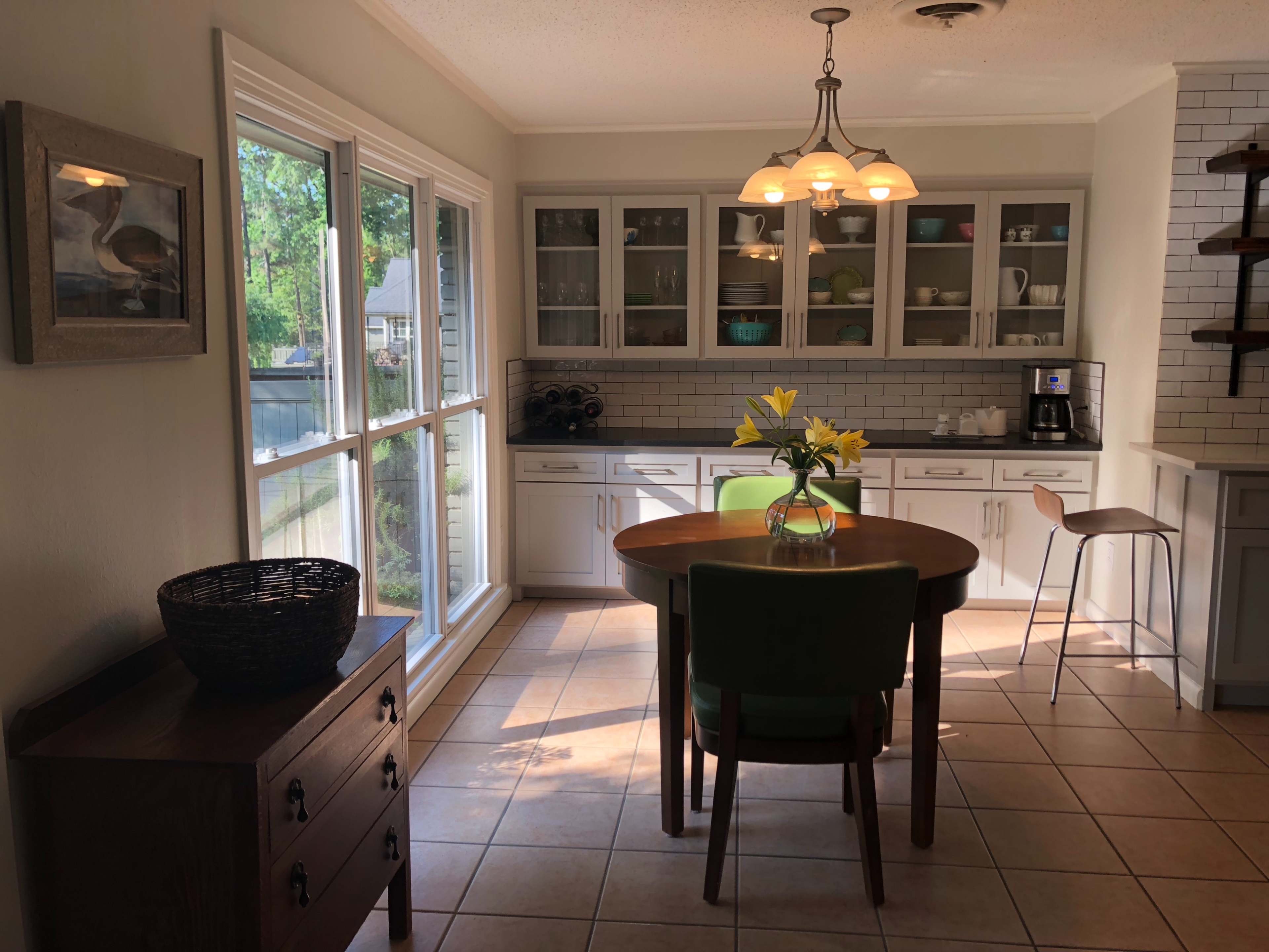 A well-lit kitchen features a round table surrounded by chairs, with a window and cabinetry in the background.