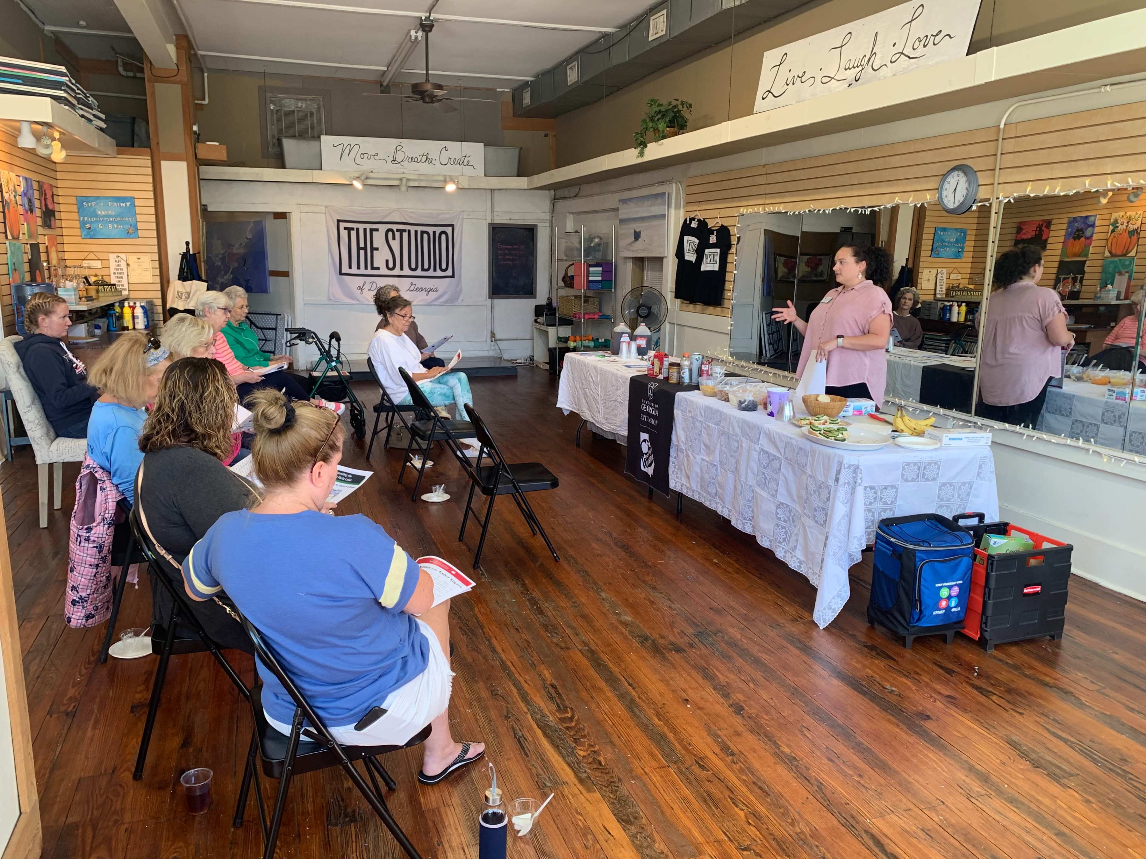 A group of people sits in chairs facing a presenter in a studio space, while a table displays various items and refreshments.